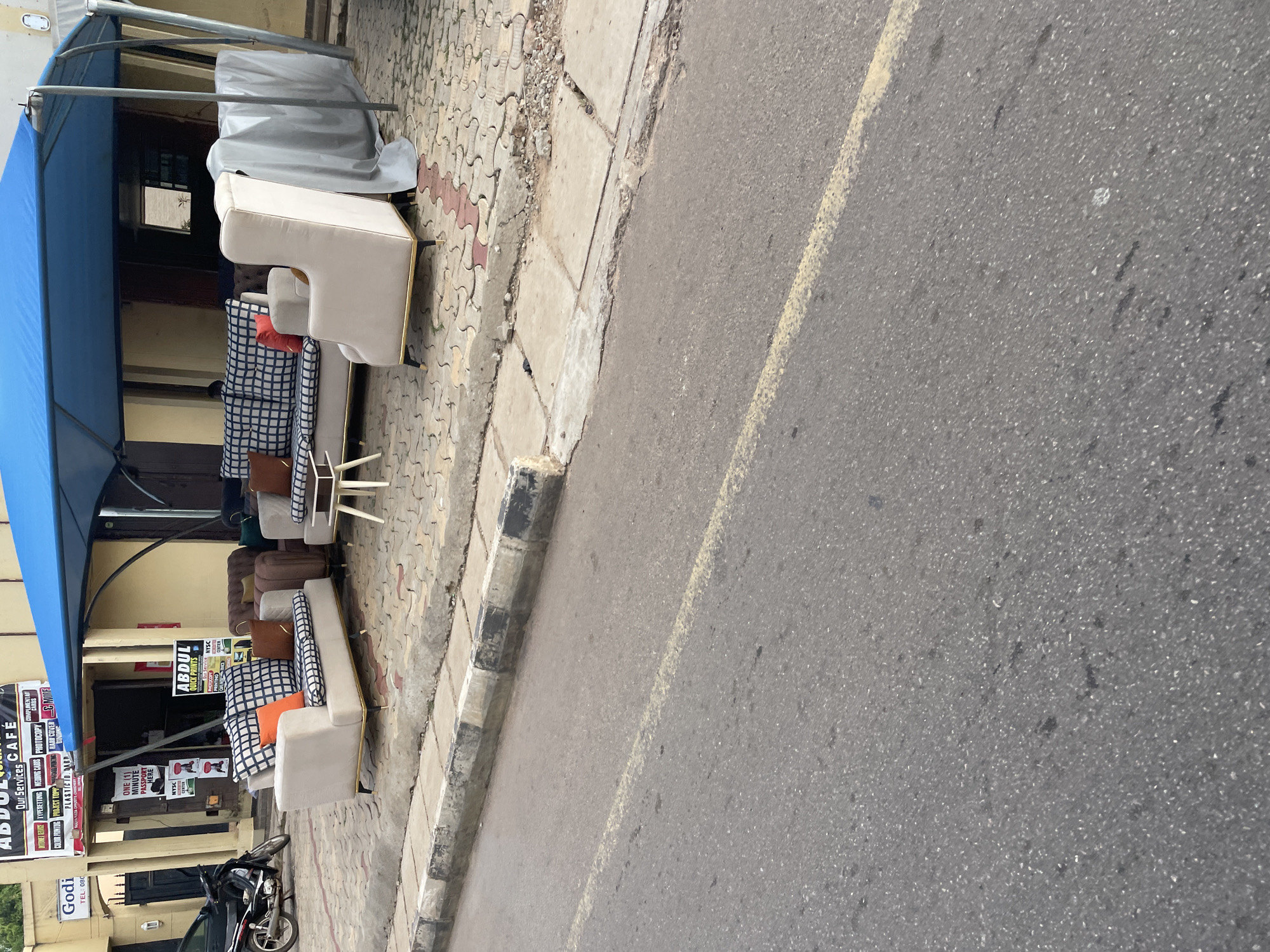 This is a photograph taken in Barden Yana, Nigeria, likely during the daytime, given the bright lighting. The image captures a street scene with a focus on furniture displayed outside a shop. A blue awning extends over the shop front, sheltering several pieces of modern furniture, including a light-colored sofa and armchairs. The furniture is arranged on a patterned paved walkway next to a street. Some of the seating features checkered and orange cushions.
Behind the furniture, the shop entrance is visible, with signs indicating "ABDUL QUICK PRINTS" and "ABDUL Our Services CAFÉ". Other text on signs includes "INTERNET VENICE TYPESETTING WEDDING CARDS PHOTOCOPY CARDS" and references to "NYSC CENTER". A motorcycle is parked on the pavement near the shop. The street itself appears to be asphalt with a faded yellow line marking. The overall impression is that of a commercial area with a furniture display extending onto the public walkway.