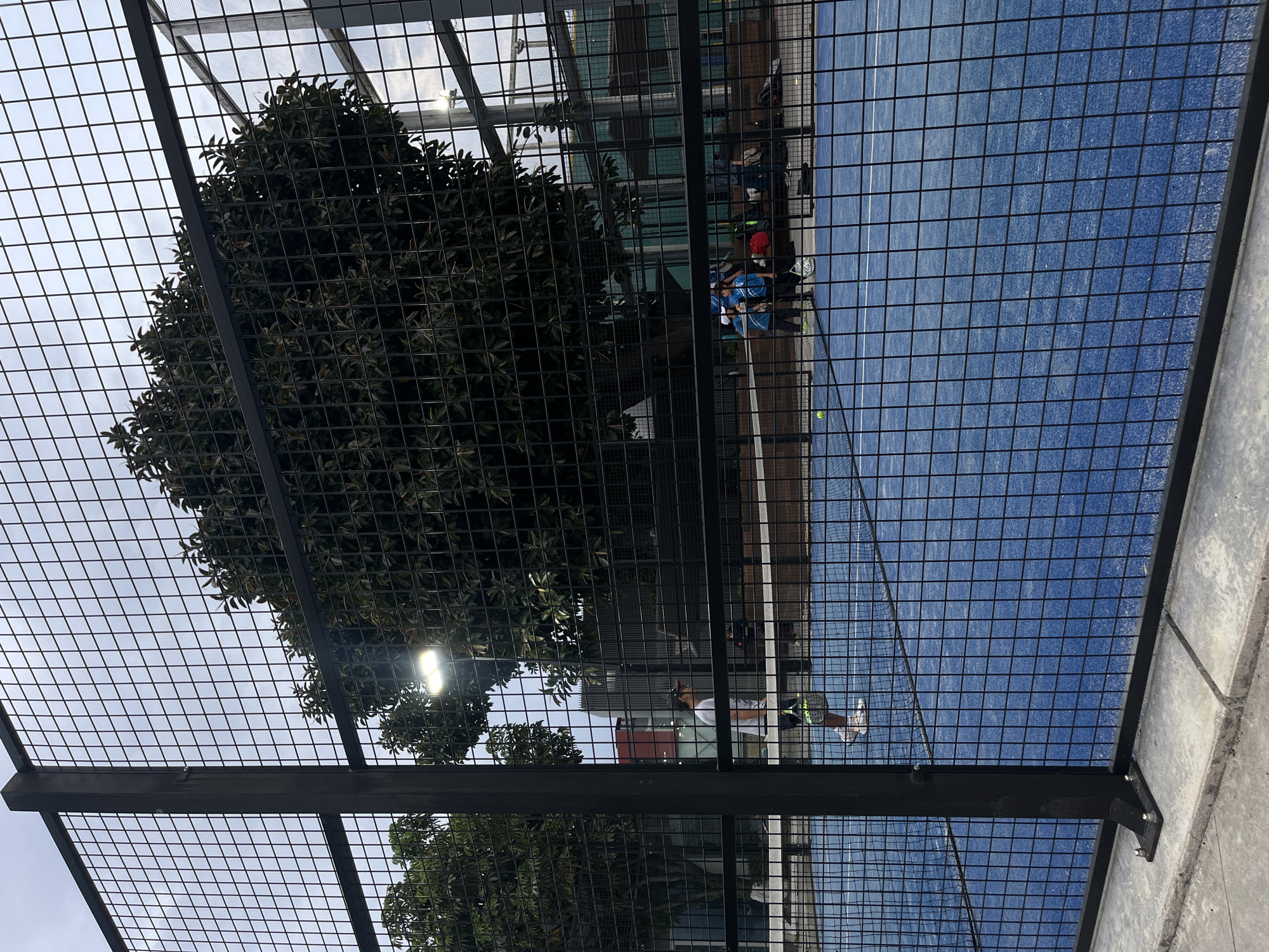 This outdoor, oblique view of a paddle tennis court is heavily obscured by a black metal wire fence. On the blue-painted court surface, a solitary individual is seen in mid-swing, presumably hitting a tennis ball. Further into the background, figures are sitting on benches. There's a tree visible through the fencing. An artificial light is also visible. The location is Unknown, Unknown.