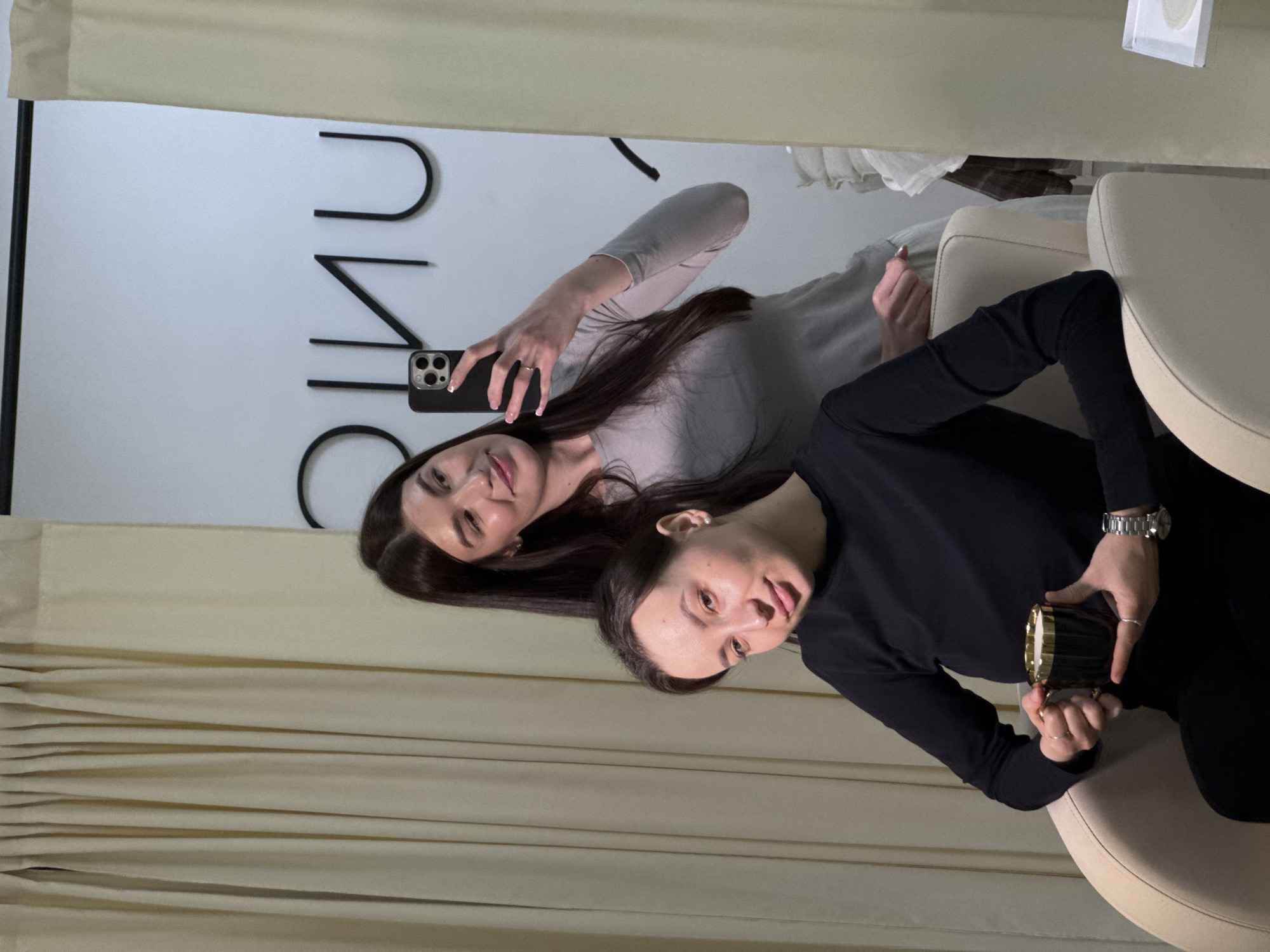 Two women are taking a selfie indoors, possibly in a shop. One woman is holding a black smartphone, likely taking the photo. She is wearing a light grey long-sleeved top. The other woman is seated in a cream-colored chair, wearing a black long-sleeved top and holding a black and gold container. A silver watch is visible on her wrist. There is a dark inscription on the wall behind them, and beige curtains frame the scene. This is possibly a hair salon or a similar establishment. The city of Chaikovsky, Russia, is not identifiable in the image without further information.
