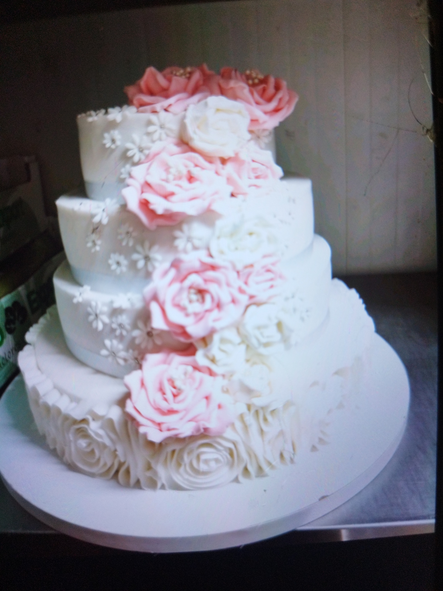 A four-tiered cake is centrally positioned on a white platform, likely made of cardboard or similar material. The cake is adorned with white and pink frosting in floral patterns. Roses, crafted from frosting, are prominent, with white blossoms and smaller daisy-like flowers sprinkled on the sides. The lowest tier has a textured, swirl-like frosting design. The cake appears to be set indoors on a reflective, metallic surface. The backdrop consists of a light-colored wall, possibly tiled. A green object is partially visible to the left of the cake. There is no indication of any specific location in Plovdiv, Bulgaria or any people or interactions in the scene.