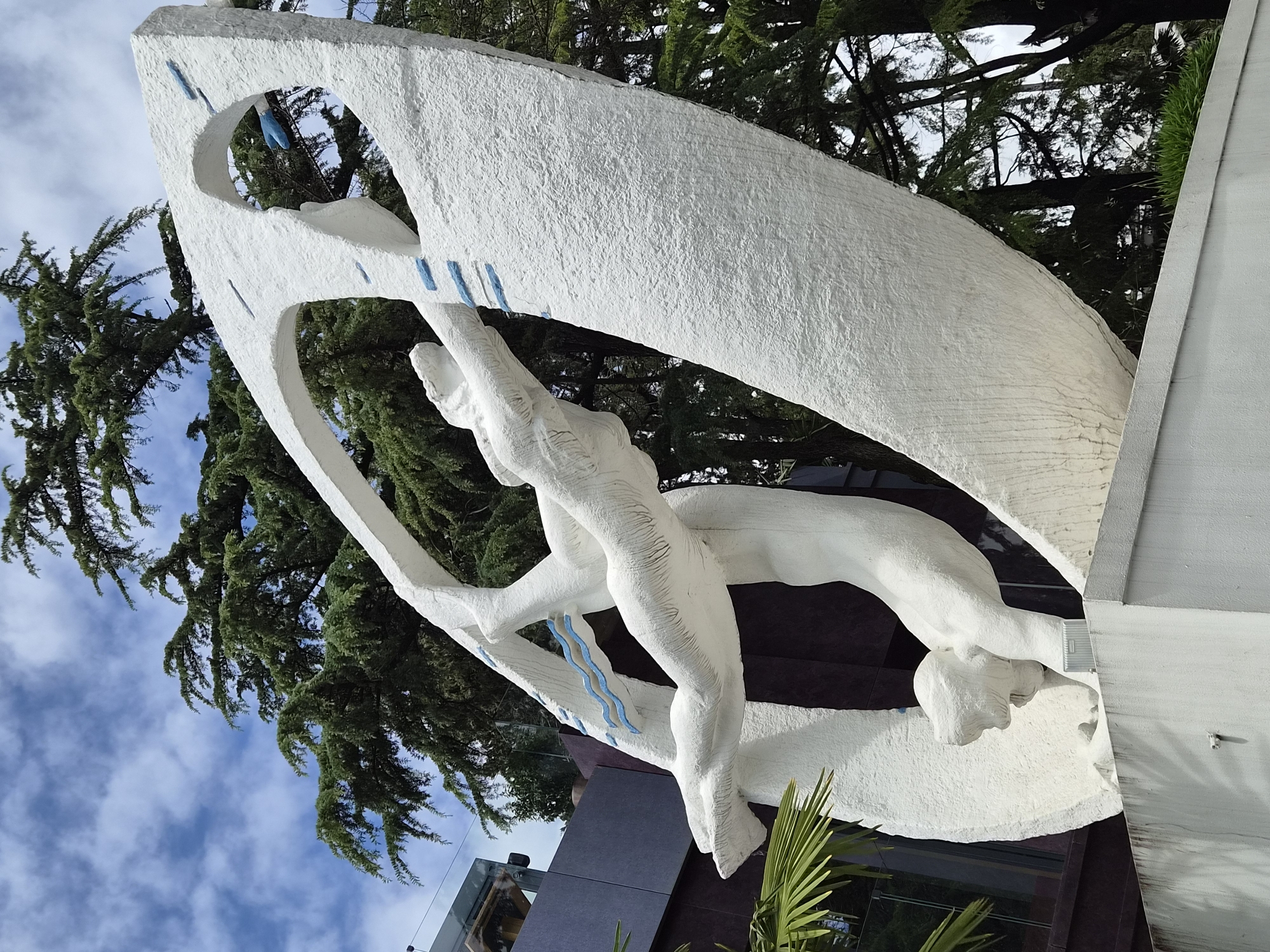 A partially obstructed, low-angle, rotated medium shot shows a large, white concrete sculpture depicting two human figures in an embrace, possibly a dance, set against a backdrop of greenery and sky. The sculpture is located in Sochi, Russia.
The sculpture features two intertwined figures, both rendered in a rough, textured white concrete. One figure appears to be supporting the other, creating a dynamic composition. The sculpture is partially framed by a large, arched white structure, also made of concrete, with geometric blue accents.
The backdrop includes dense green trees, a partly cloudy sky, and glimpses of a building with a glass façade. Vegetation is visible at the base of the structure.