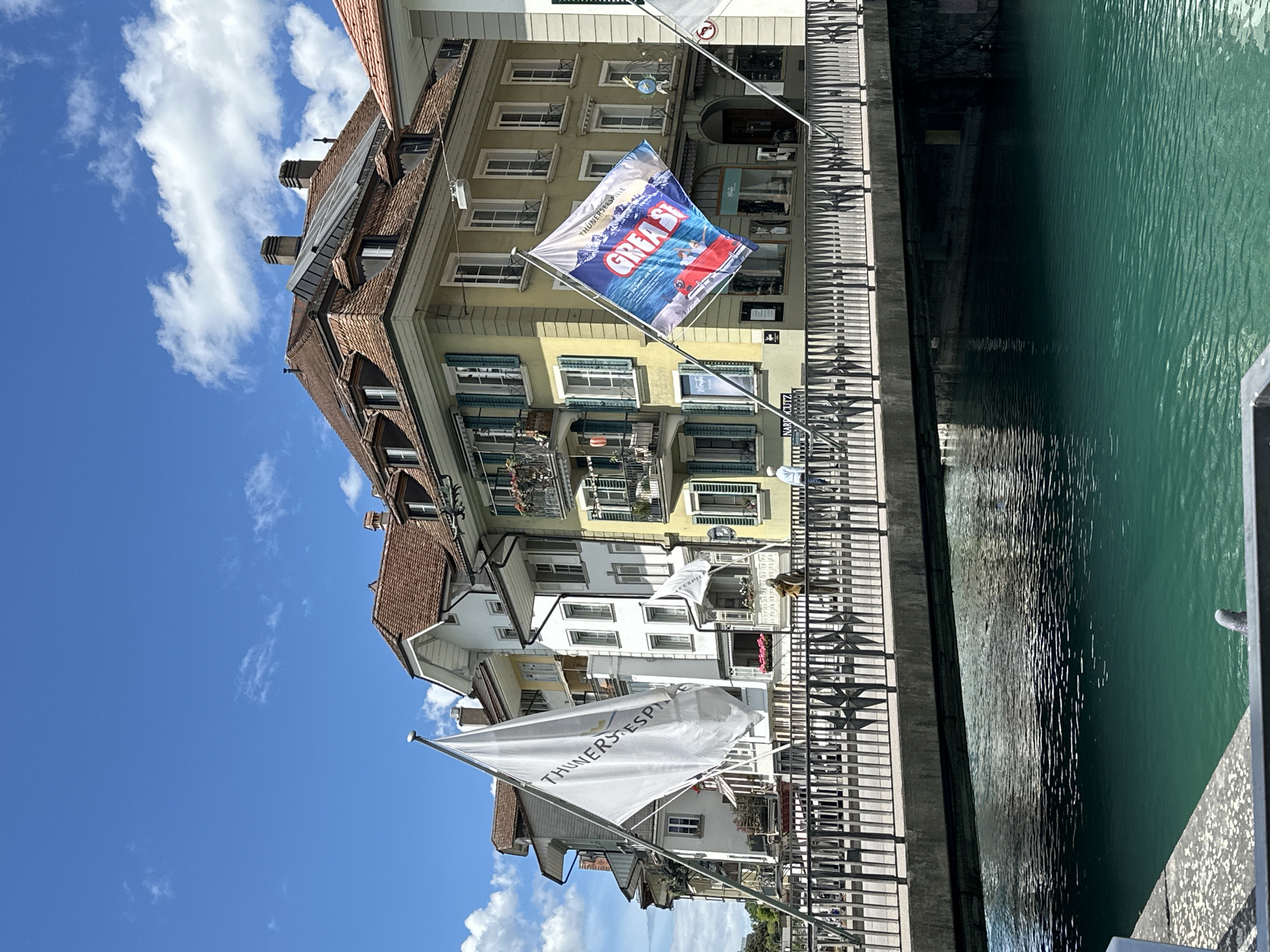 The image depicts a sunlit scene in the city of Thun, Switzerland, featuring a bridge spanning a vibrant turquoise body of water. On the bridge, two flags are prominently displayed. The flag on the left is white with black lettering that reads "THUNERSEE-SPIELE". The flag on the right is a colorful banner advertising "GREASE" with an image of a red vehicle and people. The background consists of several historic buildings with characteristic Swiss architecture, including pitched roofs, dormer windows, and colorful facades, one of which is pale yellow. The sky above is a clear blue with scattered white clouds. A few individuals are visible on the bridge and along the waterfront, suggesting a public area. The water's surface is rippled, reflecting the light and the surrounding structures.