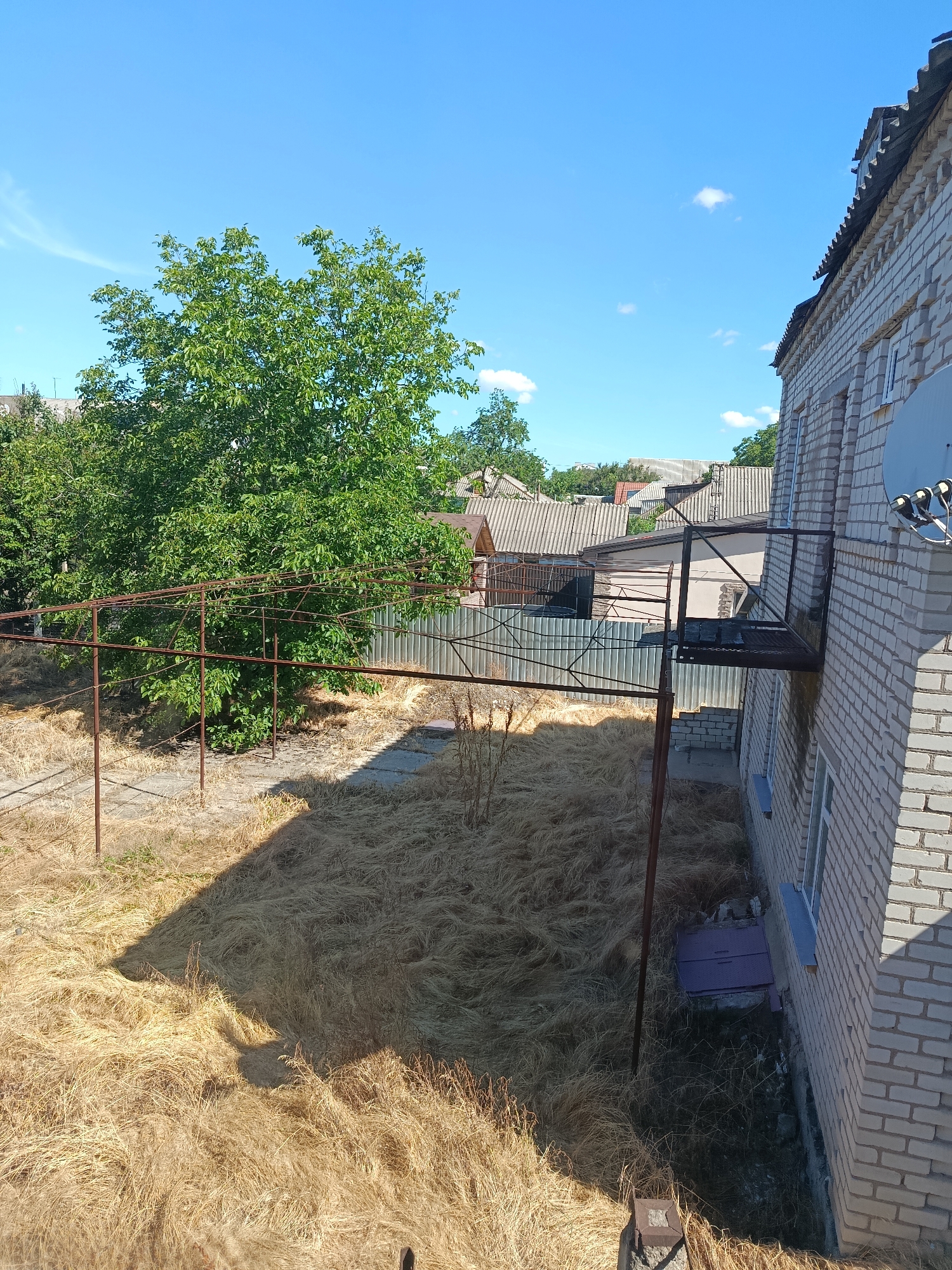 The image shows an outdoor scene on a sunny day, likely in a rural or suburban area. The foreground is dominated by dry, golden grass, suggesting a dry season or a well-used yard. A metal structure, possibly a clothesline or a trellis, stands in the middle ground, casting a shadow across the grass. To the right, a light-colored brick building with a dark roof runs along the edge of the frame. A small, dark metal balcony juts out from the building, and a satellite dish is mounted on the wall. In the background, several other buildings with grey, corrugated roofs are visible, along with a large, leafy green tree. The sky is a clear, bright blue with a few wispy white clouds. The overall impression is one of a simple, functional outdoor space, possibly a backyard. There are no people visible in the image. The time of day appears to be midday, given the bright sunlight and the distinct shadows. The weather is clear and warm.