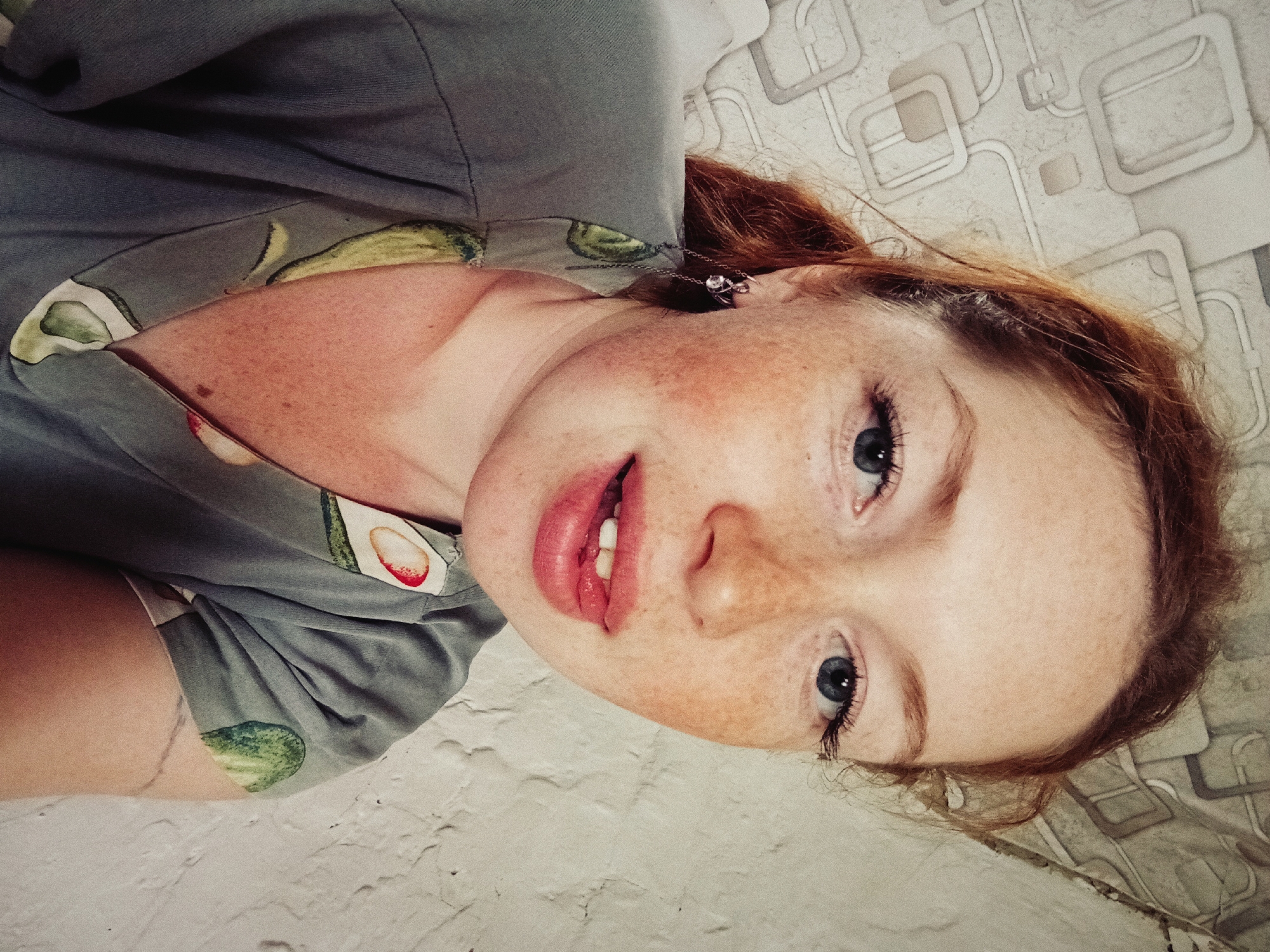 A fair-skinned, young, freckled woman with reddish-brown hair is pictured in a close-up selfie. Her eyes are blue. She is wearing a gray t-shirt with avocado prints. A silver necklace is visible on her neck. The background appears to be a wall with a textured, patterned design. The location is not discernible as Yurgamysh, Russia, based solely on the visual information. The woman's expression is neutral with her mouth slightly open. The angle of the photo is from above, looking down at the subject.