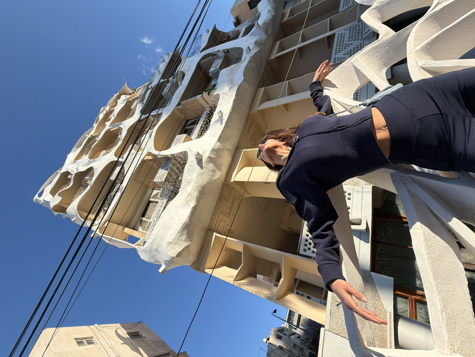 A low-angle, wide shot captures an urban scene in Tel Aviv, Israel, dominated by the distinctive Pagoda House (Beit HaPagoda) on Nahmani Street 48.
In the foreground, an adult female is positioned behind a textured, light-colored concrete railing with an irregular, wavy pattern. She wears a dark navy blue cropped, ribbed, zippered hoodie, revealing her midriff, and matching high-waisted leggings with a V-shaped seam. Her long, dark hair is down, and black sunglasses rest on her head. A light-colored shoulder bag is visible on her right side. She is captured with her head tilted sharply upwards, looking towards the sky, arms outstretched and slightly bent at the elbows, palms open, suggesting a posture of exhilaration or surrender. A ring is visible on her left ring finger.
The background is dominated by the Pagoda House, a multi-story building characterized by its organic, undulating, white concrete facade that forms wave-like structures around balconies and windows. The building exhibits intricate metalwork railings on its balconies and some external window blinds. Below the main white facade, parts of the building feature a tan/beige color scheme with subtle geometric patterns etched into the surface. To the far left, another simpler, light beige building is partially visible. The sky above is a clear, vibrant blue, and multiple dark electrical wires stretch horizontally across the upper frame. The scene is illuminated by strong, natural sunlight, casting distinct shadows.