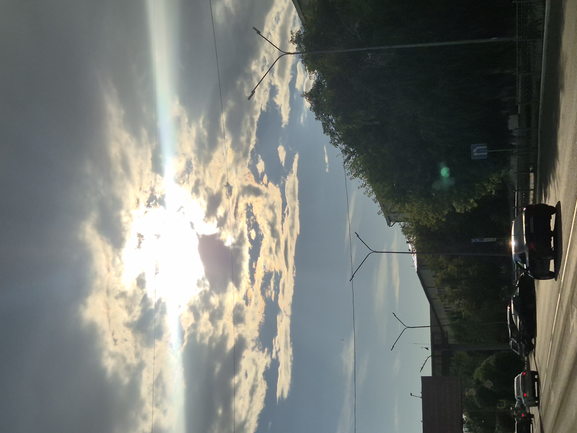 The image captures a view of a street in Kokshetau, Kazakhstan, with cars driving on a multi-lane road. The sky is dramatic, with bright sunlight piercing through scattered, dark clouds, creating lens flare effects. On the right side of the road, there are tall trees and buildings, along with streetlights. On the left, another building and a large sign are visible. The perspective is from within a vehicle, looking forward.