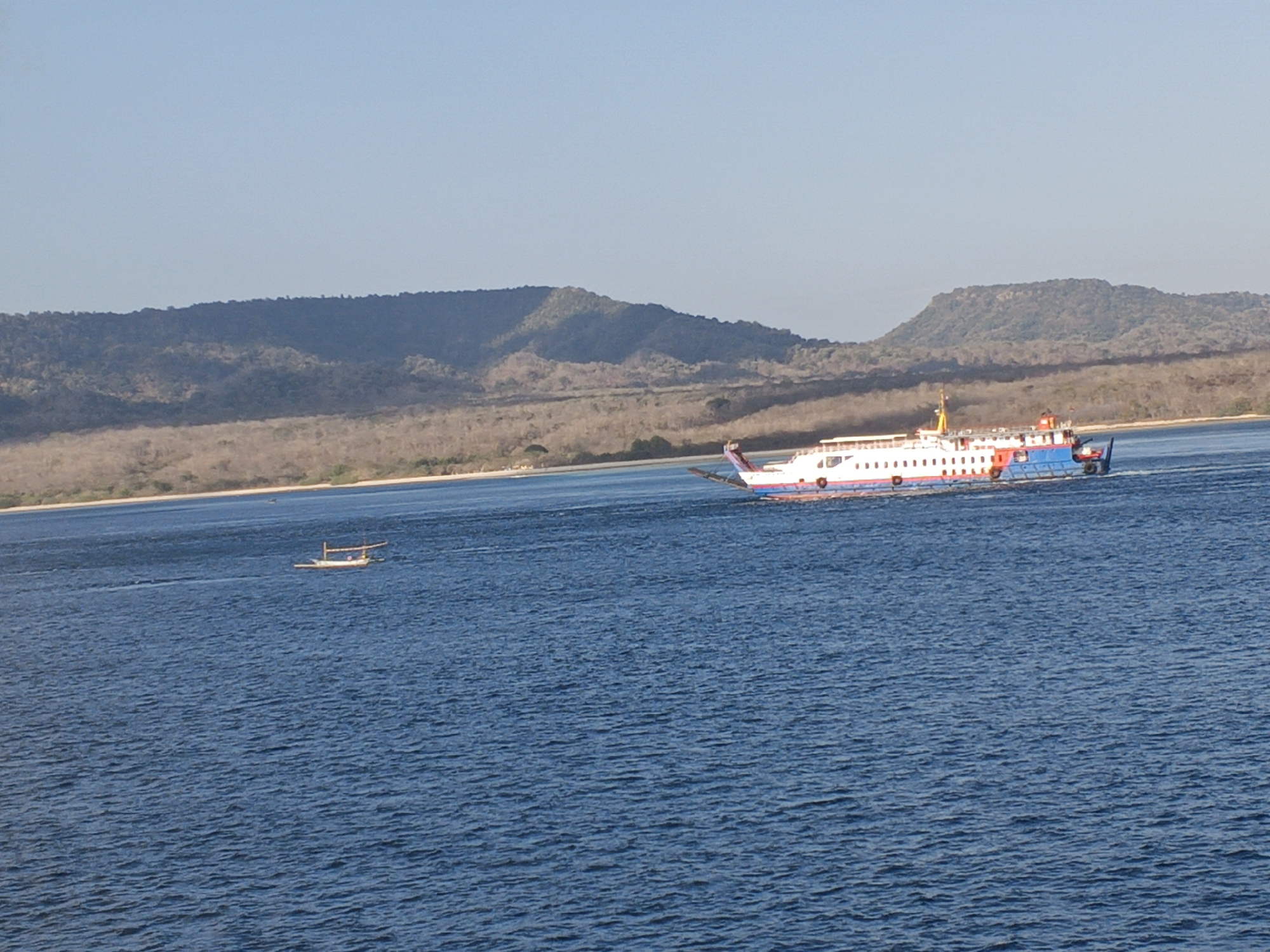 A wide shot captures the ocean near Melaya, Indonesia, with a large ferry, the "KM Dharma Kencana I," navigating the waters. The ferry, predominantly white with red and blue accents, has numerous windows and appears to be a passenger vessel. In the distance, a smaller, traditional wooden boat with an outrigger is visible. The background features a coastline with dry, undulating hills covered in sparse vegetation. The sky is clear and a pale blue, suggesting a sunny day. The water is a deep blue with subtle ripples.