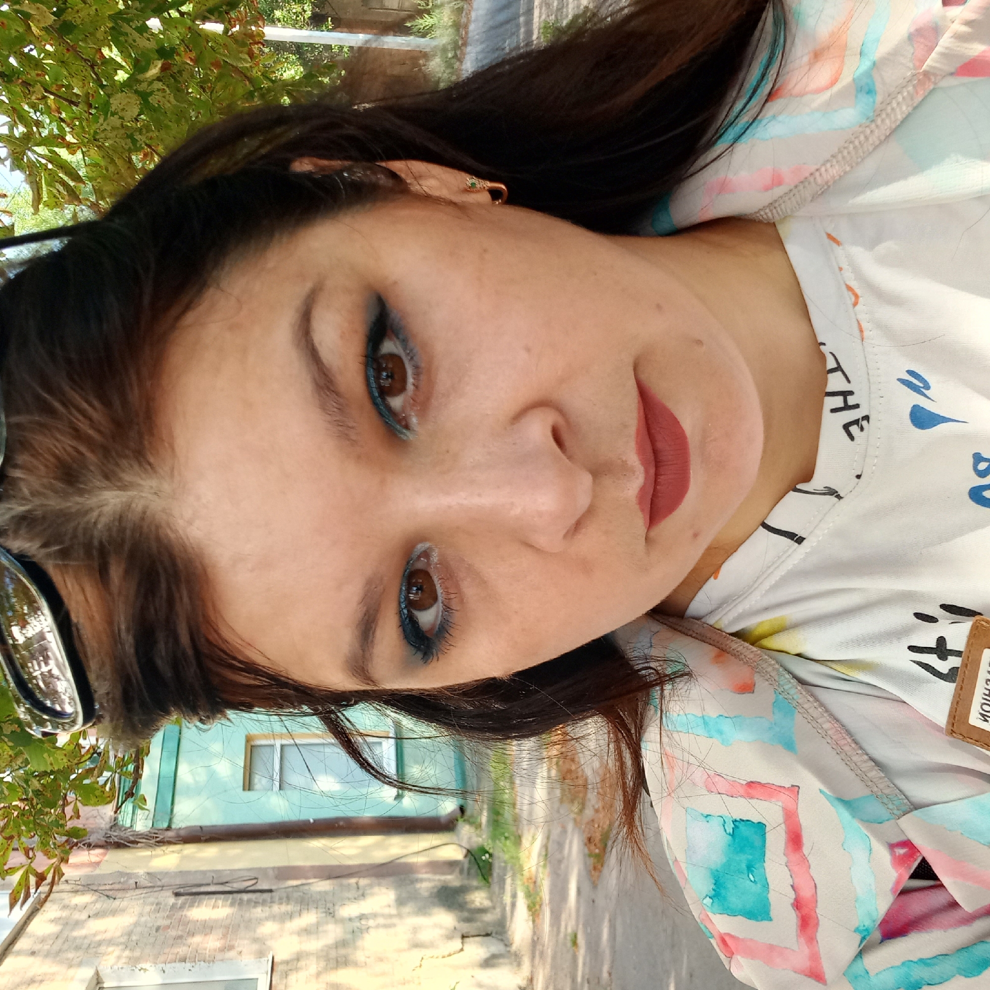 A close-up, waist-up, outdoor selfie captures a woman with medium-toned skin and dark brown hair. She is wearing a white t-shirt with black text and a colorful patterned jacket over it. Her eyes are adorned with dark blue eyeshadow and eyeliner. Her lips are painted with a matte reddish-brown lipstick. The background shows a street scene with buildings, one of which is painted a light turquoise, and trees with green and yellowing leaves. The lighting suggests it is daytime. The location is Kropyvnytskyi, Ukraine.