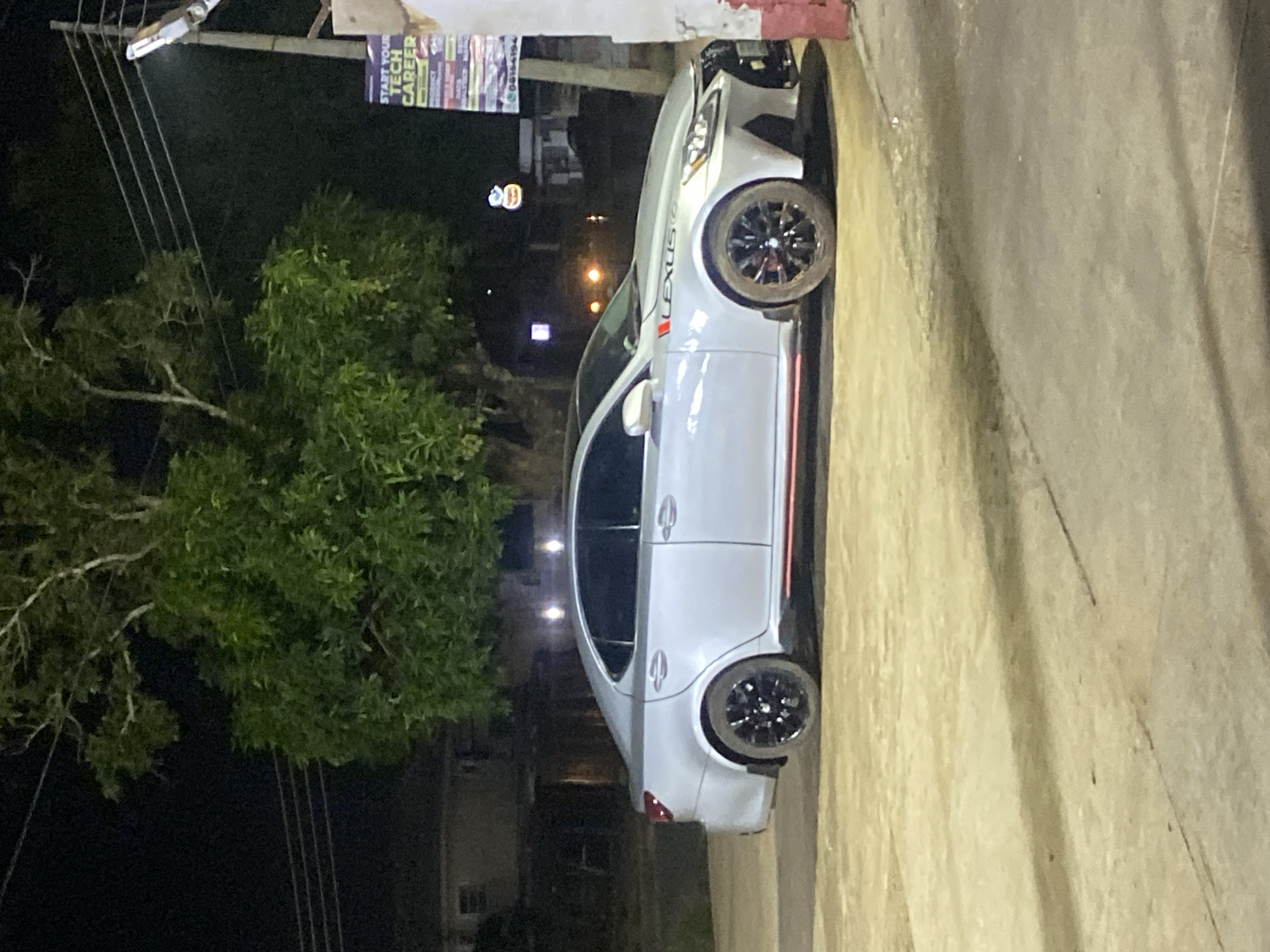 The image depicts a static nighttime scene in Ikorodu, Nigeria, centered on a light silver Lexus sedan parked on a dusty, light-colored ground surface. The car, possibly an IS model, features black multi-spoke wheels, which are visibly soiled with reddish-brown dirt or mud, particularly on the front passenger wheel. Red accent stripes are present on the lower side skirts and a smaller vertical stripe accompanies the "LEXUS" lettering on the front fender. The car's upper body, including the roof and area above the door handles, appears to be a darker grey or black, contrasting with the lighter silver lower body.
In the background, a large tree with dense green foliage dominates the upper center of the frame. To the left, a dark building with faint window outlines is visible. To the right, a utility pole supports multiple power lines and an illuminated street light fixture. A partially visible commercial sign, positioned vertically, displays the text "START YOUR TECH CAREER." A reddish-brown wall or structure is present on the extreme right, partially overlapping the car's front. In the distant background, blurred lights and an illuminated, distinct logo are visible. No individuals are present, and no actions or interactions are depicted.