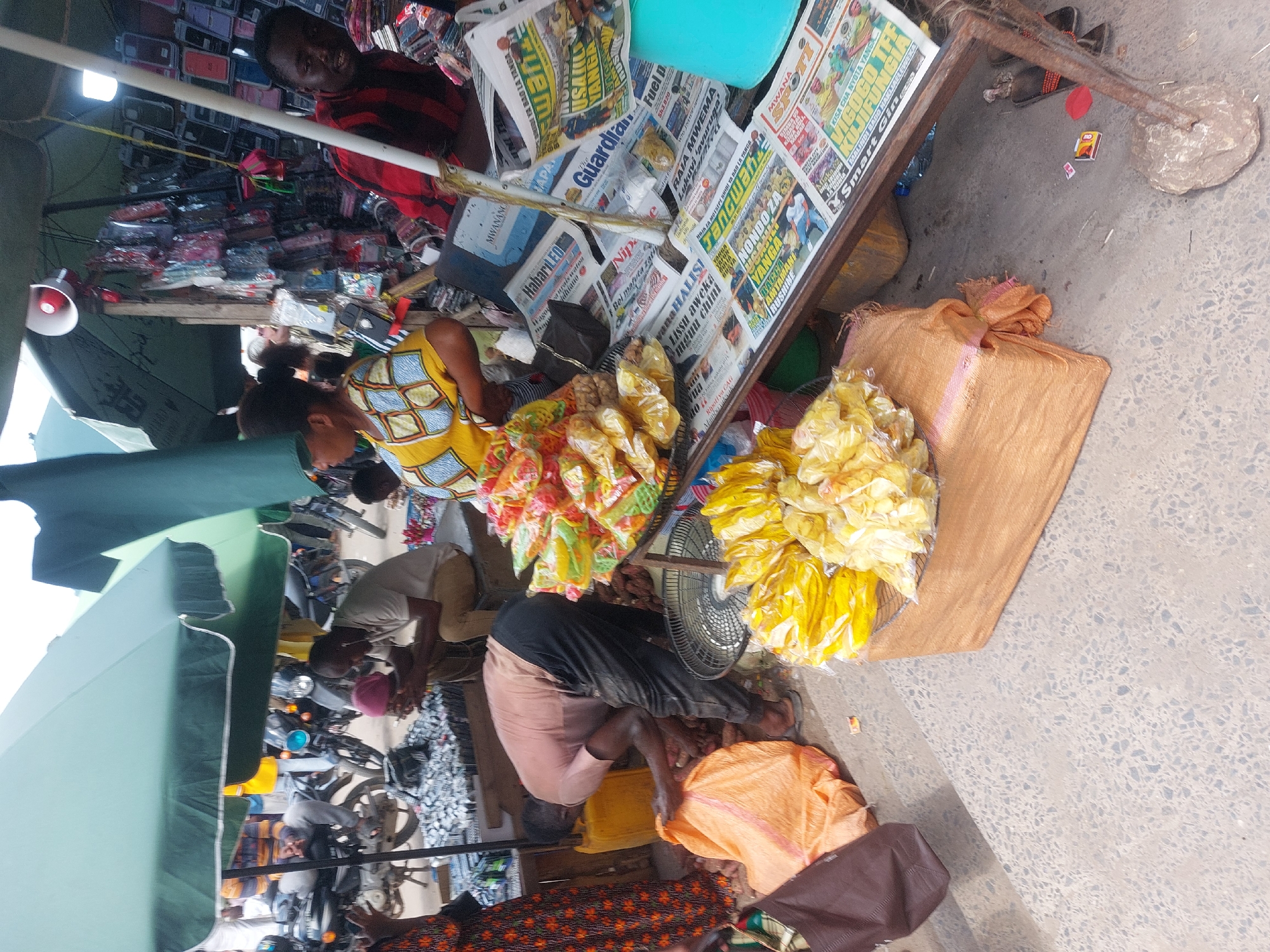 The image depicts an outdoor scene in what appears to be a bustling market or roadside stall in Dar es Salaam, Tanzania. A woman with a yellow and blue patterned top is tending to two heaps of packaged snacks; one is a multicolored, yellow, orange, and green package, the other a predominantly yellow package, and are arranged on a table covered with newspapers. The newspapers feature headlines and stories in Swahili, the national language of Tanzania. A portable electric fan sits near the yellow packages. A man in a red striped shirt is at a stall with multiple cell phone cases.
Several people are milling around. One man is kneeling and appears to be sorting items on the ground. Other people are visible in the background, including people on motorbikes. A large green umbrella provides shade to the people. The ground is concrete, and there's a collection of motorbikes. The overall impression is that of a vibrant, busy, and somewhat chaotic commercial environment.