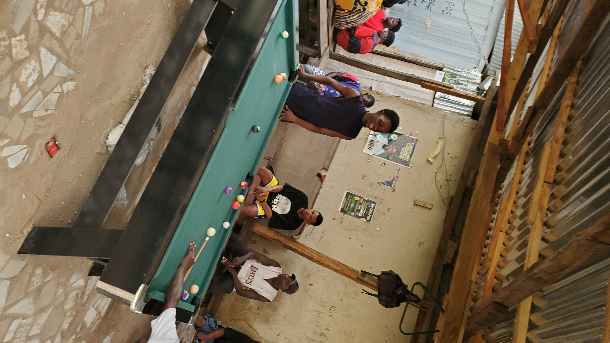 The image depicts an outdoor pool hall in Jalingo, Nigeria, characterized by a tin roof supported by wooden beams, creating a rustic, open-air ambiance. Several individuals are present, with the foreground dominated by a green pool table. A young man in a dark sleeveless shirt is actively engaged in playing pool, leaning over the table with a cue stick in hand, seemingly about to strike a ball. Another man in a black t-shirt with yellow shorts is seated to the right of the pool table, observing the game. Further to the right, a man in a white Lakers jersey and a cap is also seated, looking towards the table. In the background, other individuals are visible, some seated on benches and one standing. The environment appears to be a casual gathering spot, with walls adorned with posters and debris on the ground. The overall scene suggests a moment of leisure and recreation.