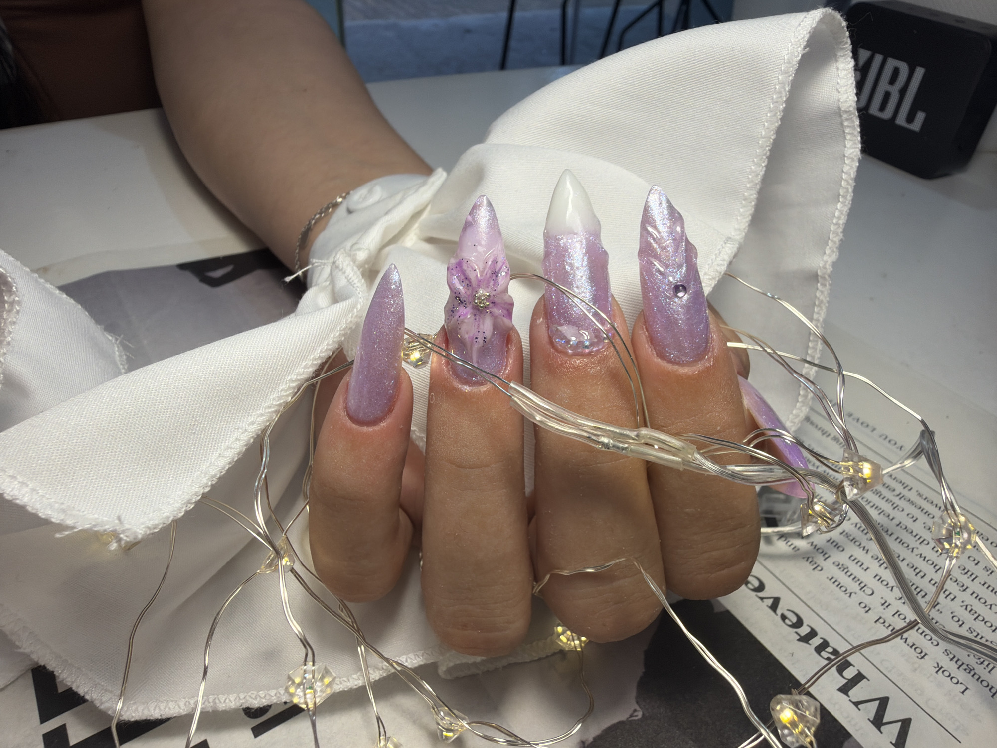 An overhead close-up shot captures a light-skinned hand, likely female, with a silver chain bracelet visible on the wrist. Five fingers display elaborately manicured artificial nails, all long and pointed. The nails are primarily a shimmering light purple/lavender color. The middle finger nail features a three-dimensional floral design in shades of purple and white, accented with a small central rhinestone. The ring finger nail presents a white tip transitioning into the purple base, with small clear embellishments near the cuticle. The pinky and index finger nails exhibit textured patterns, possibly feather-like, each with a small embedded rhinestone. The thumb nail is partially visible, also in shimmering purple.
Delicate string lights, composed of thin silver wires and small, clear, faceted bulbs, are draped across the fingers and a piece of folded white fabric. Several of these bulbs emit a warm yellow light. The white fabric, possibly a napkin or towel, shows visible stitching along its edges and partially covers a light-colored surface.
In the upper right background, a black JBL brand speaker with its logo visible is present. Below the speaker and the white fabric, a newspaper or magazine page is visible, displaying large black text, with the word "Whatever" clearly legible. The scene is situated in an indoor environment in Stara Zagora, Bulgaria.
