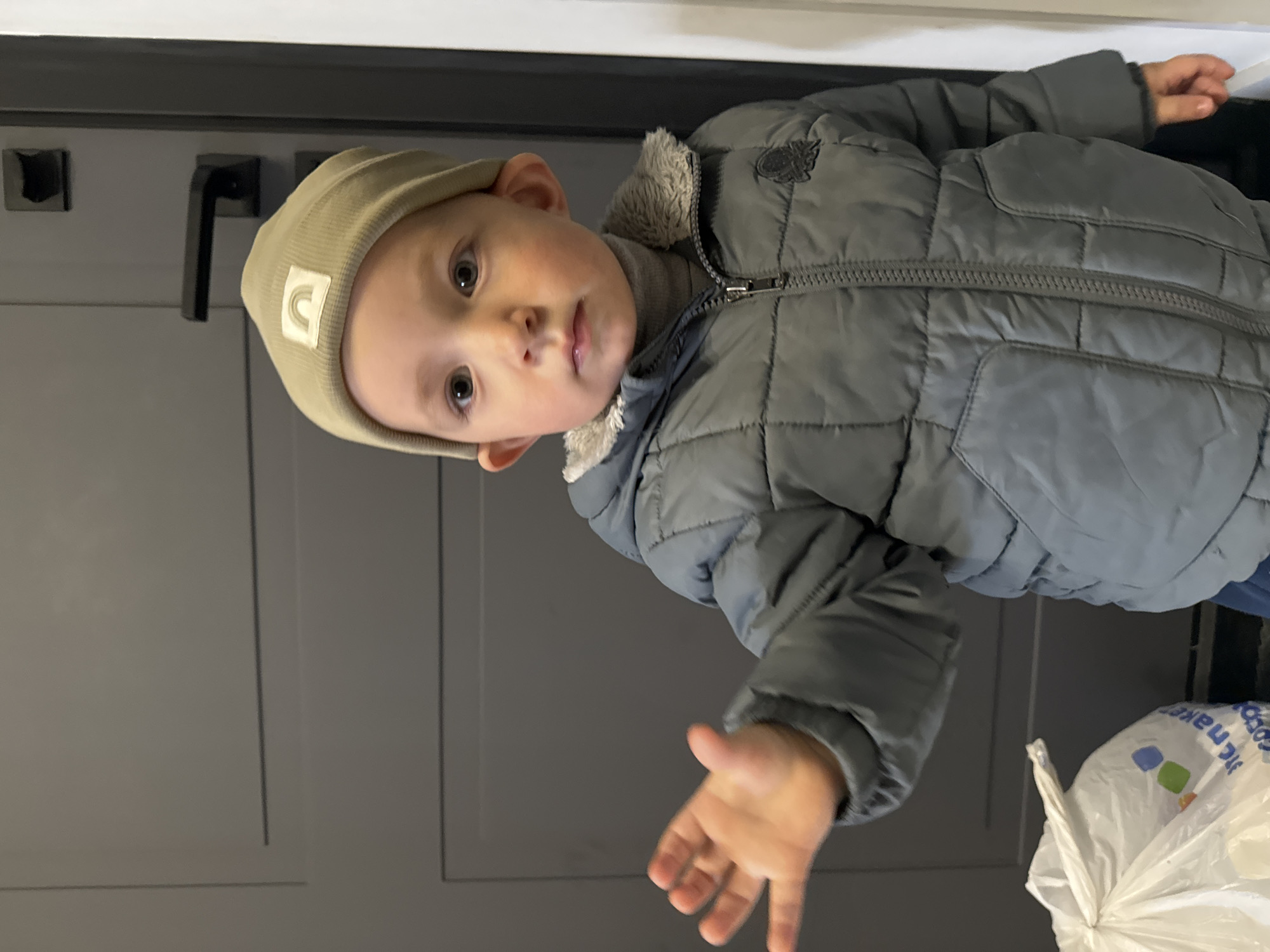 A young child wearing a gray quilted jacket and a beige hat stands in front of a gray door. The child has their right hand extended towards the viewer. A white plastic bag with printed text is visible in the lower right corner of the frame. The scene appears to be indoors, likely within a residence. The location is unspecified and not identifiable from the image provided. No specific information is available to identify that this scene is in the city of Odintsovo, Russia.