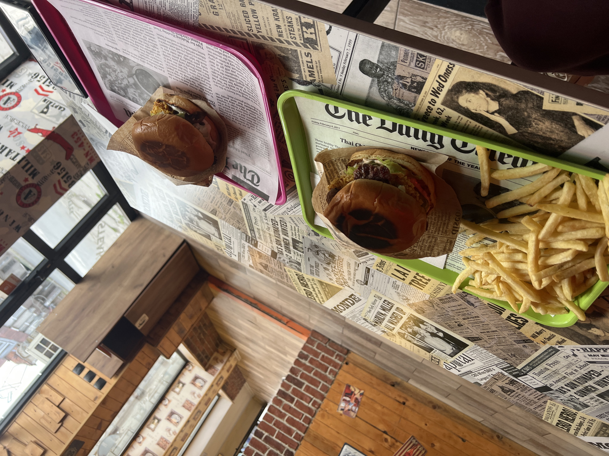 The image shows a table in a restaurant, covered with newspaper-print paper. On the table are two trays. One is pink and holds a burger wrapped in paper. The other tray is green and holds a burger and a portion of french fries. The environment shows the interior of a restaurant with wood paneling, brick elements, and a window. The location in Yambol, Bulgaria cannot be determined solely from the image.