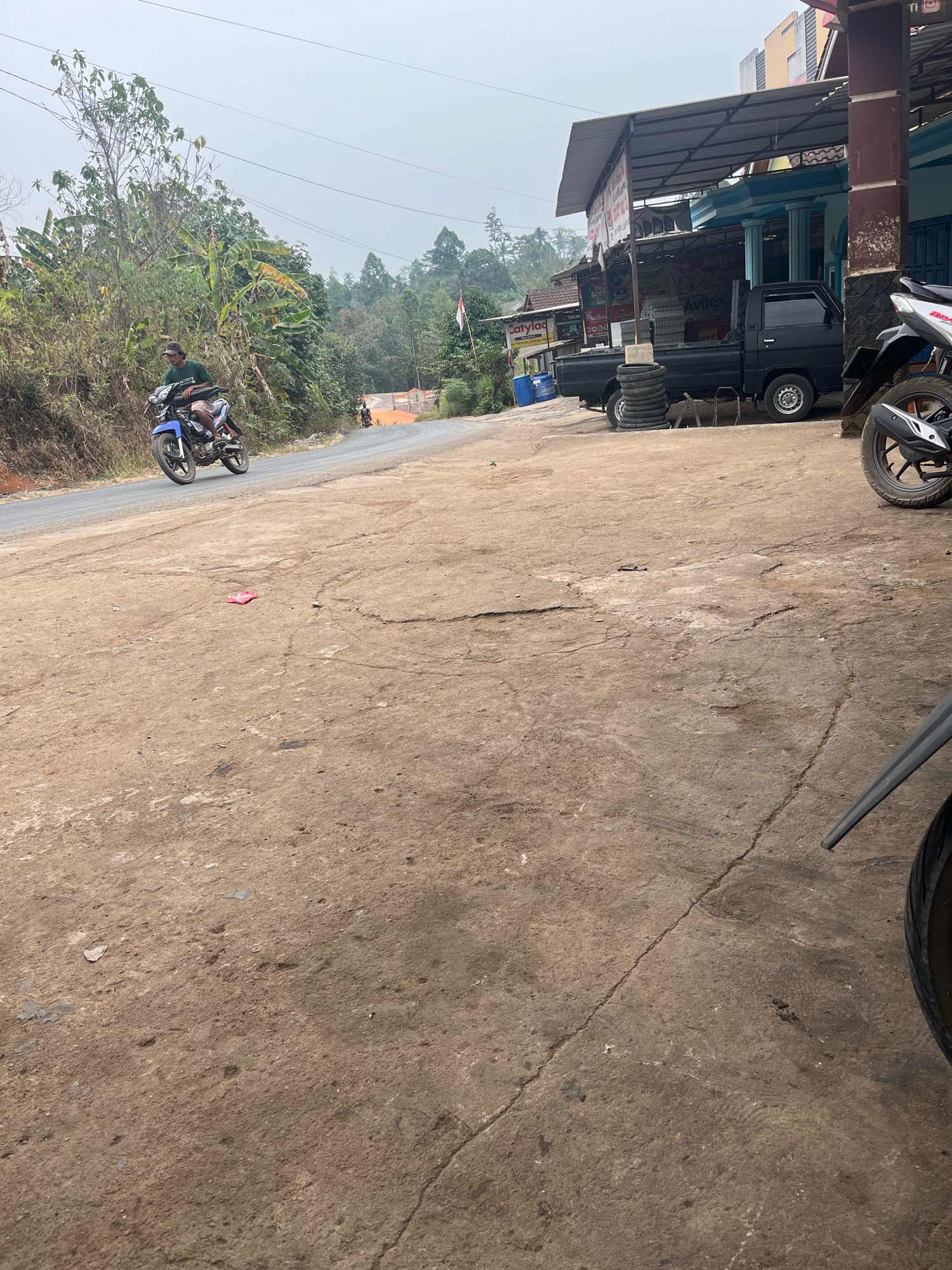 A man rides a blue motorcycle on a paved road in Karang Sari, Indonesia. The man wears a dark green t-shirt, checkered shorts, and a baseball cap. He is leaning slightly forward as he navigates the road. In the background, a black pickup truck is parked in front of a building with a blue facade and a corrugated metal roof. The building appears to be a storefront, with signage visible. To the left of the road, lush vegetation and trees line a hillside. The sky is overcast. The road surface is a mix of asphalt and dirt, with some cracks and debris visible.