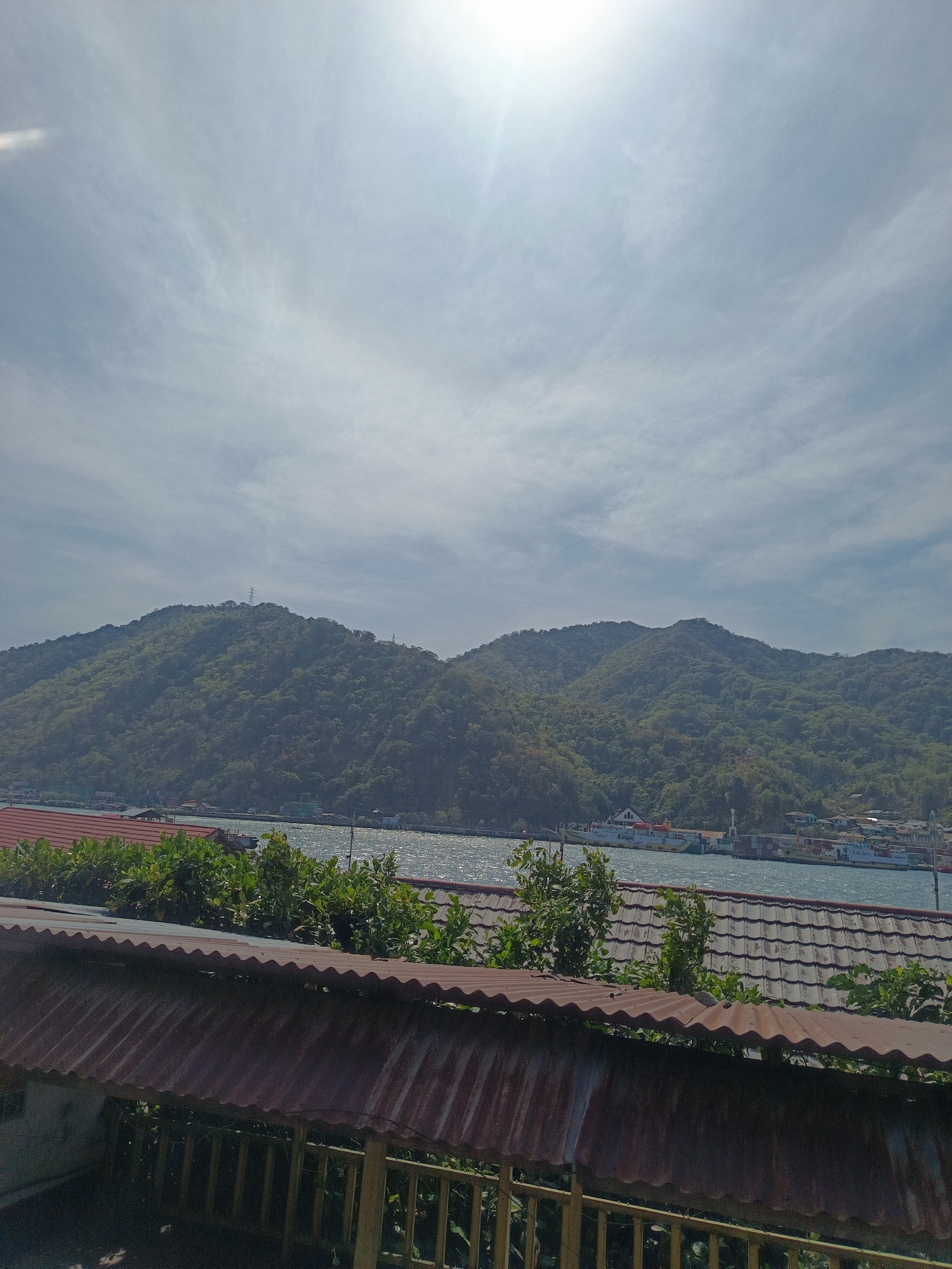 The image shows a panoramic view of a coastal area with lush green hills in the background and a body of water in the middle ground. In the foreground, there is a structure with a rusted corrugated metal roof and wooden railings. Beyond this structure, there are more buildings with red tiled roofs and dense green foliage. The water is shimmering with sunlight, and several cargo ships and smaller vessels are docked along the shoreline. The sky is bright with scattered clouds, and the sun is partially visible at the top of the frame. The scene appears to be a bustling port or harbor area in a tropical or subtropical region, likely in Gorontalo, Indonesia, given the presence of cargo ships and the characteristic hilly terrain adjacent to the sea.
