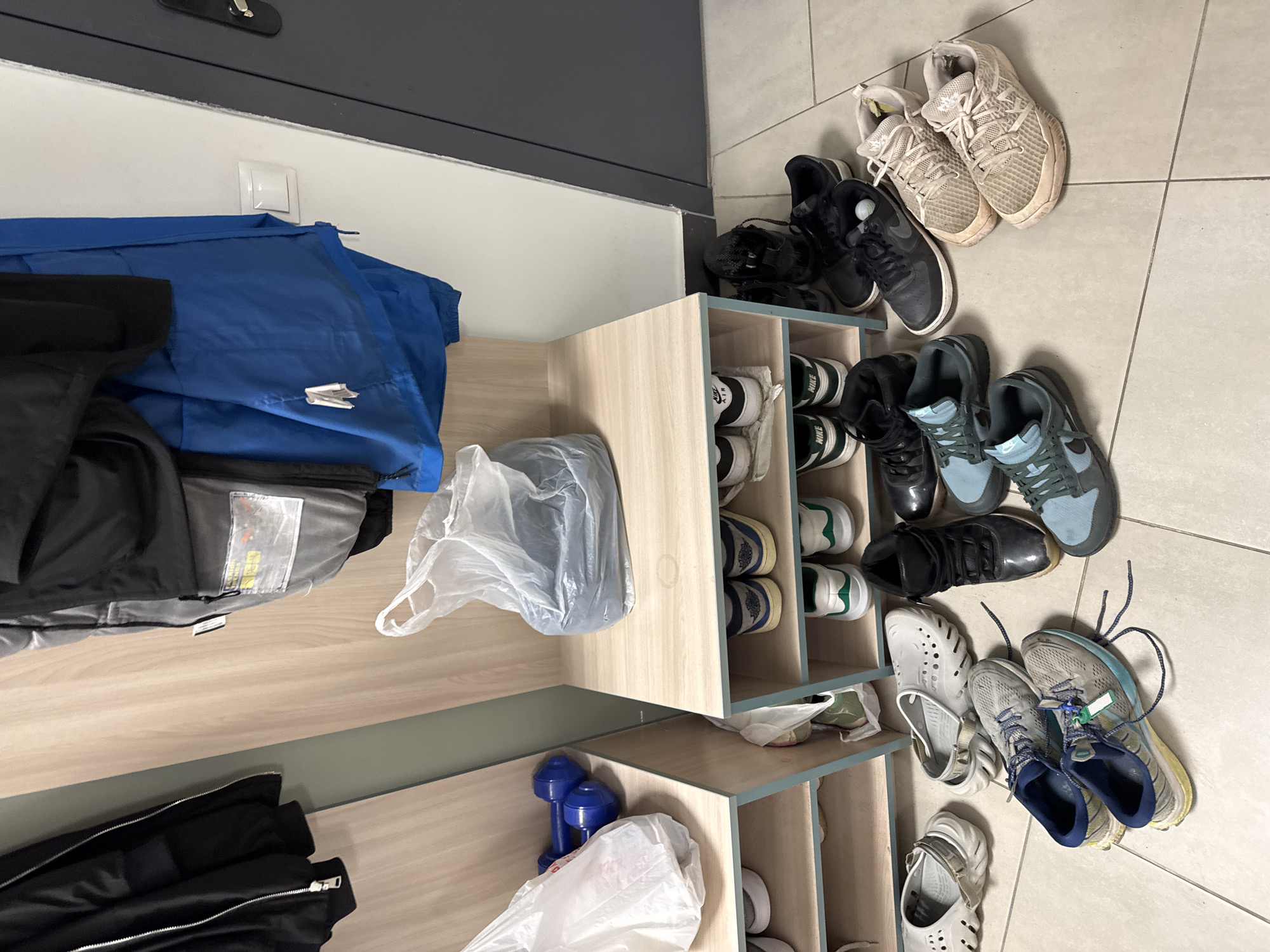 The scene appears to be an indoor entryway or mudroom. A wooden storage unit is visible, containing items such as shoes, a pair of blue dumbbells, and clothing. A collection of shoes is scattered on the tiled floor adjacent to the storage unit. A blue jacket and other black clothes lie adjacent to the wooden storage unit. There is a light switch on the wall. The specific location in the city of Kazan, Russia, is not discernible from the image.