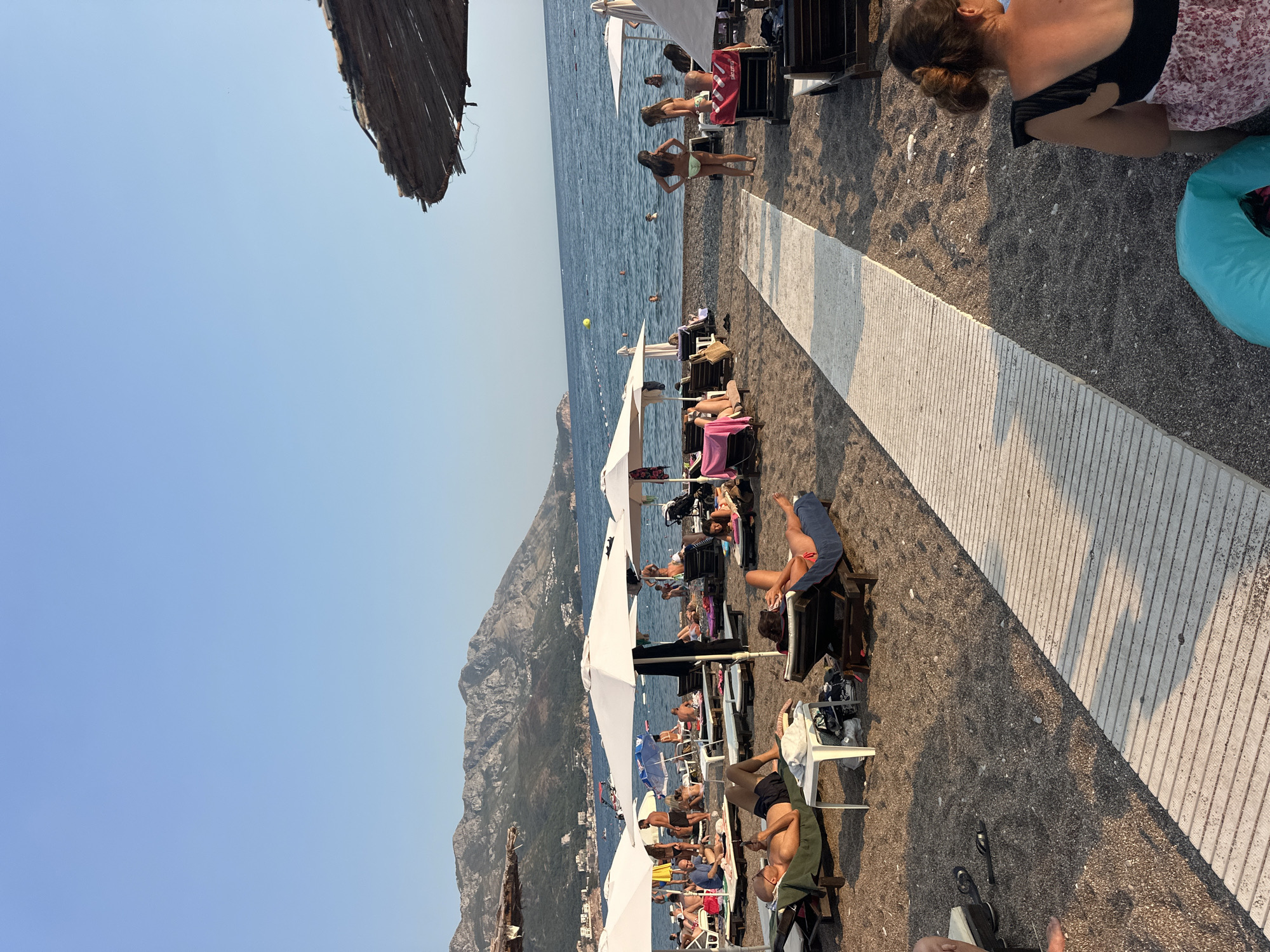 The image depicts a crowded beach scene in Boreti, Montenegro. The beach is covered with dark grey pebbles and sand. In the foreground, a white wooden walkway stretches diagonally across the frame. Several beach chairs with people relaxing on them are placed on either side of the walkway. White umbrellas provide shade for some of the chairs.
In the mid-ground, people are visible in the calm blue sea, some swimming and others wading. Further out, the sea meets a hazy horizon with distant mountains and buildings. The sky is clear and blue. In the background, a thatched umbrella is visible in the upper right corner. People are scattered throughout the scene, engaging in various beach activities, mostly lounging on chairs. The overall atmosphere is one of leisure and relaxation on a sunny day.