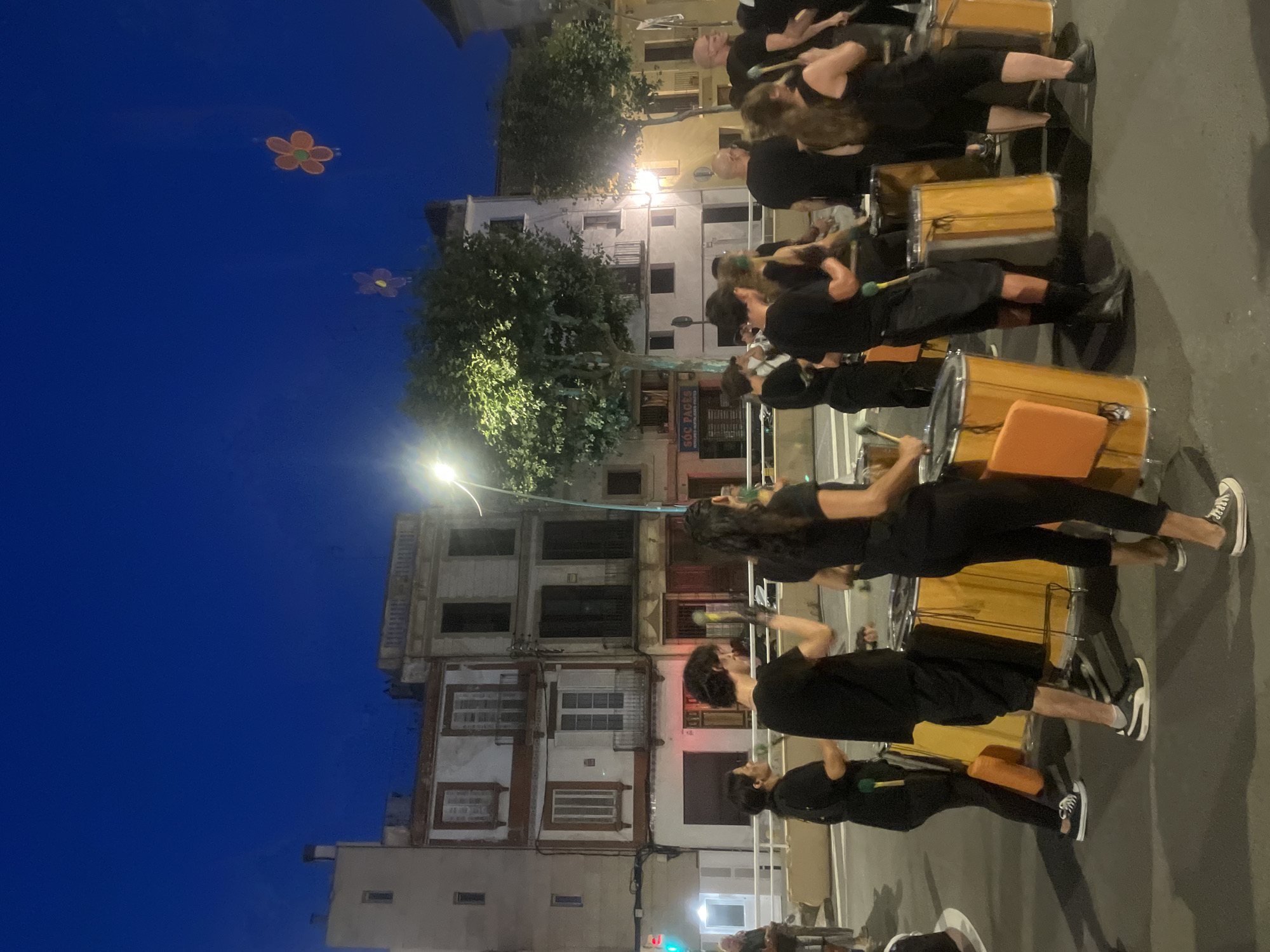 In the city of Arenys De Mar, Spain, at dusk, a group of individuals in black attire are seen playing drums. The scene is set on a street with buildings in the background. Two illuminated flower-shaped lanterns are visible in the dark blue sky. The people are positioned in front of a row of large, cylindrical drums, some of which are orange. They are holding drumsticks and appear to be in the midst of a performance or parade. The environment suggests an outdoor event or celebration.