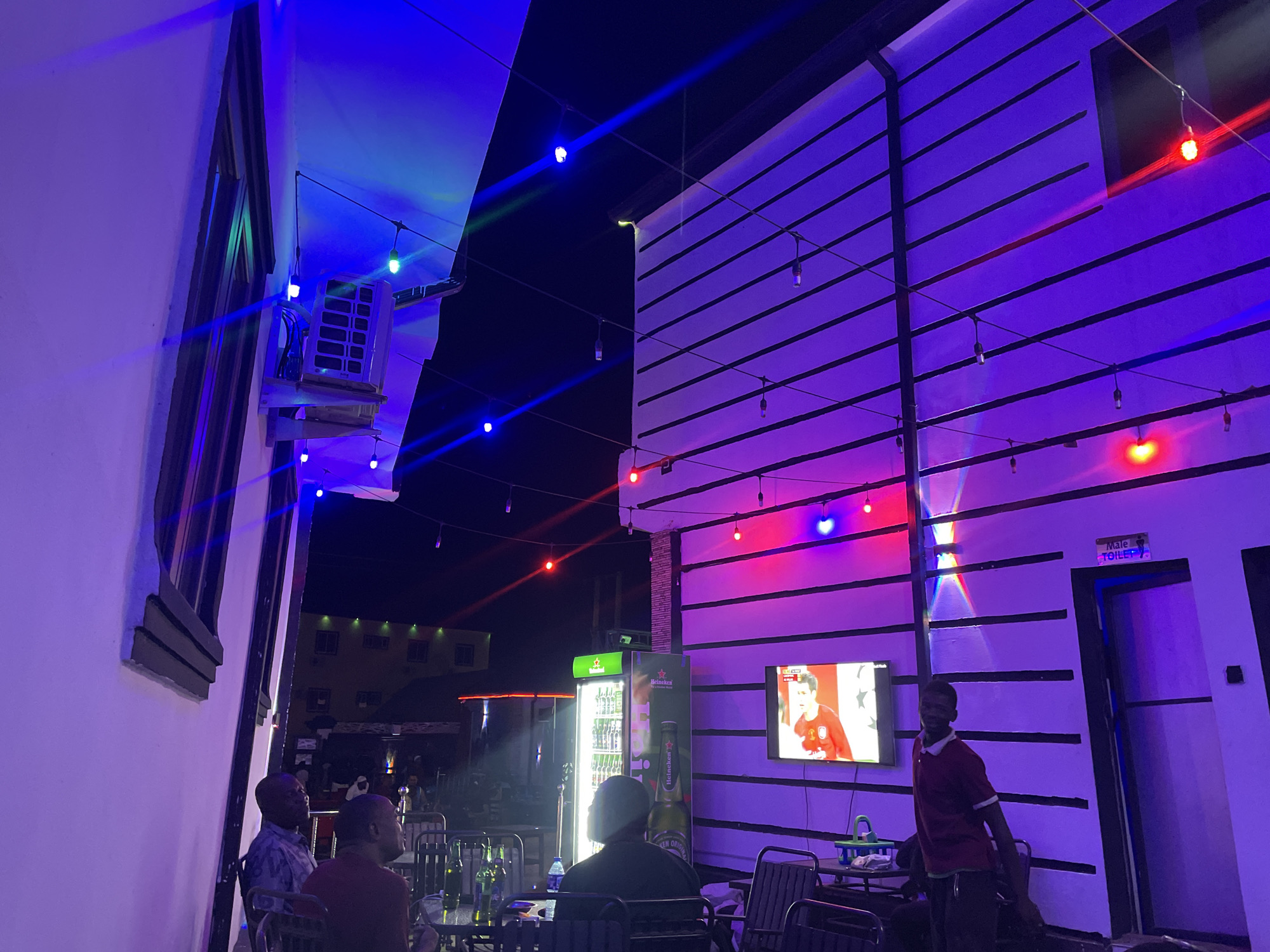 The image depicts an outdoor bar or lounge in Tumfure, Nigeria, at night. The scene is illuminated by string lights of various colors, including blue and red, and strong ambient blue and purple lighting.
Several people are visible. Three men are seated at a table to the left, appearing to be engaged in conversation. A man wearing a red shirt is standing to the right, seemingly working at the bar.
Objects in the scene include tables, chairs, beer bottles, a tall refrigerator stocked with beer, and a television mounted on the wall, displaying a soccer match. A sign indicating "Male Toilet" is visible above a doorway. The architectural style of the building is characterized by white walls with black trim and horizontal bands.