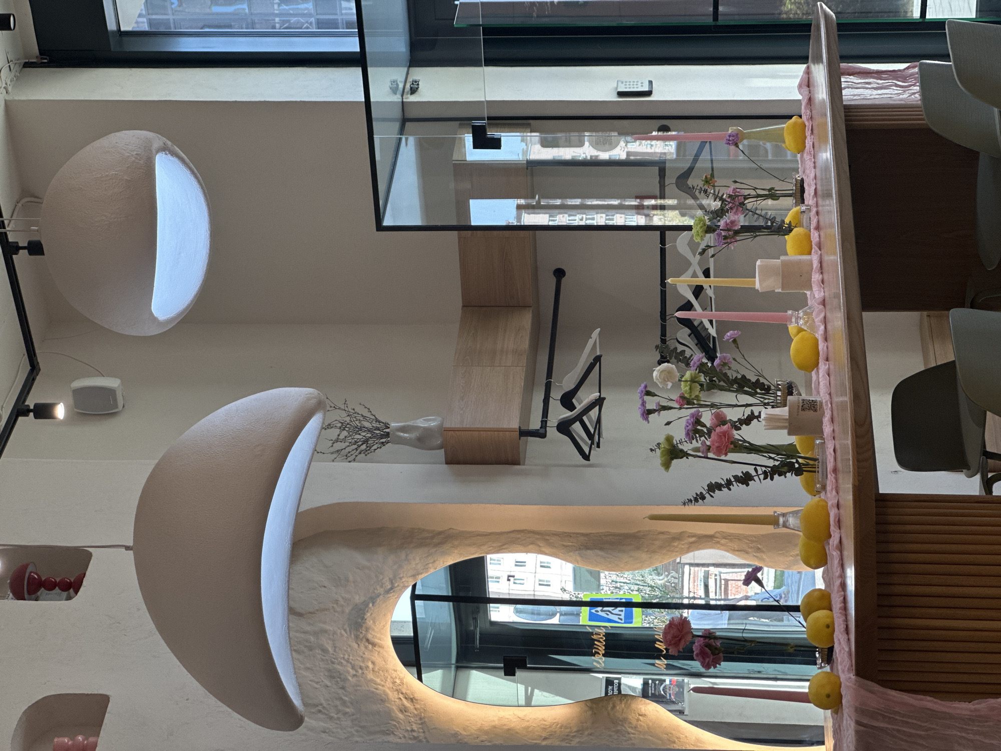 The image shows the interior of a cafe or restaurant. A prominent feature is a curved, illuminated design element in the wall, resembling a crescent moon. There is also a spherical lighting fixture on the ceiling. A table with floral decorations, candles, and lemons is visible. In the background, a window reflects the urban landscape, possibly showing buildings and a street. The presence of signage with Cyrillic script, combined with the street view, suggests that the location might be in Novosibirsk, Russia.