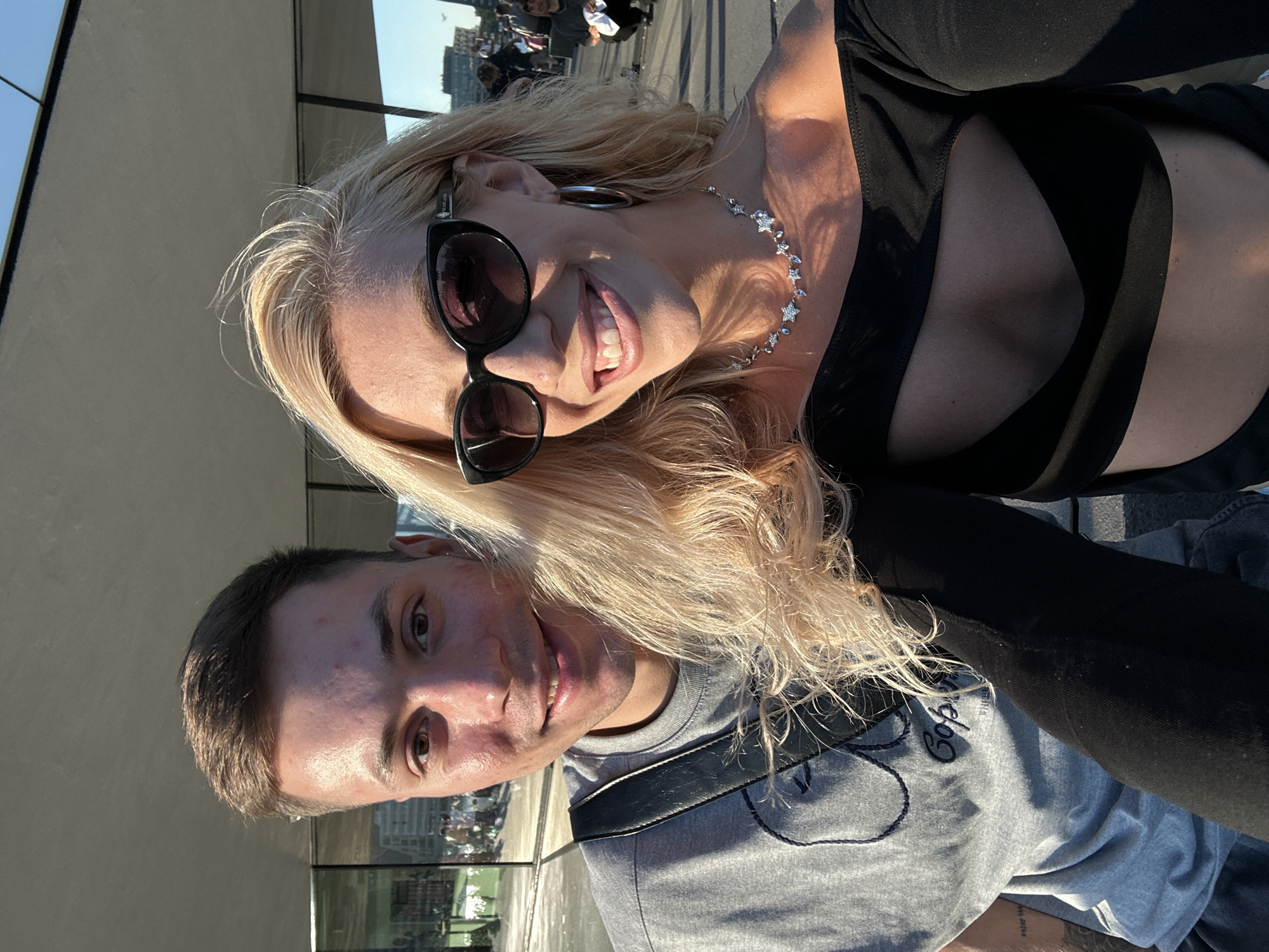A close-up, selfie-style photograph captures a couple outdoors, appearing to be at the Oeverpark in Amsterdam, Netherlands. The man, positioned on the left, wears a gray t-shirt with a dark emblem and a black crossbody bag strap. He has short brown hair and is smiling. The woman, on the right, has blonde hair, wears black sunglasses, large hoop earrings, and a black off-the-shoulder top with a cutout. She is also smiling and wearing a silver necklace with star and crystal pendants. The background suggests an urban setting with a modern architectural structure, glass facades, and a glimpse of the sky. The lighting indicates it is daytime, likely late afternoon or early evening, given the warm, golden hue of the sunlight.