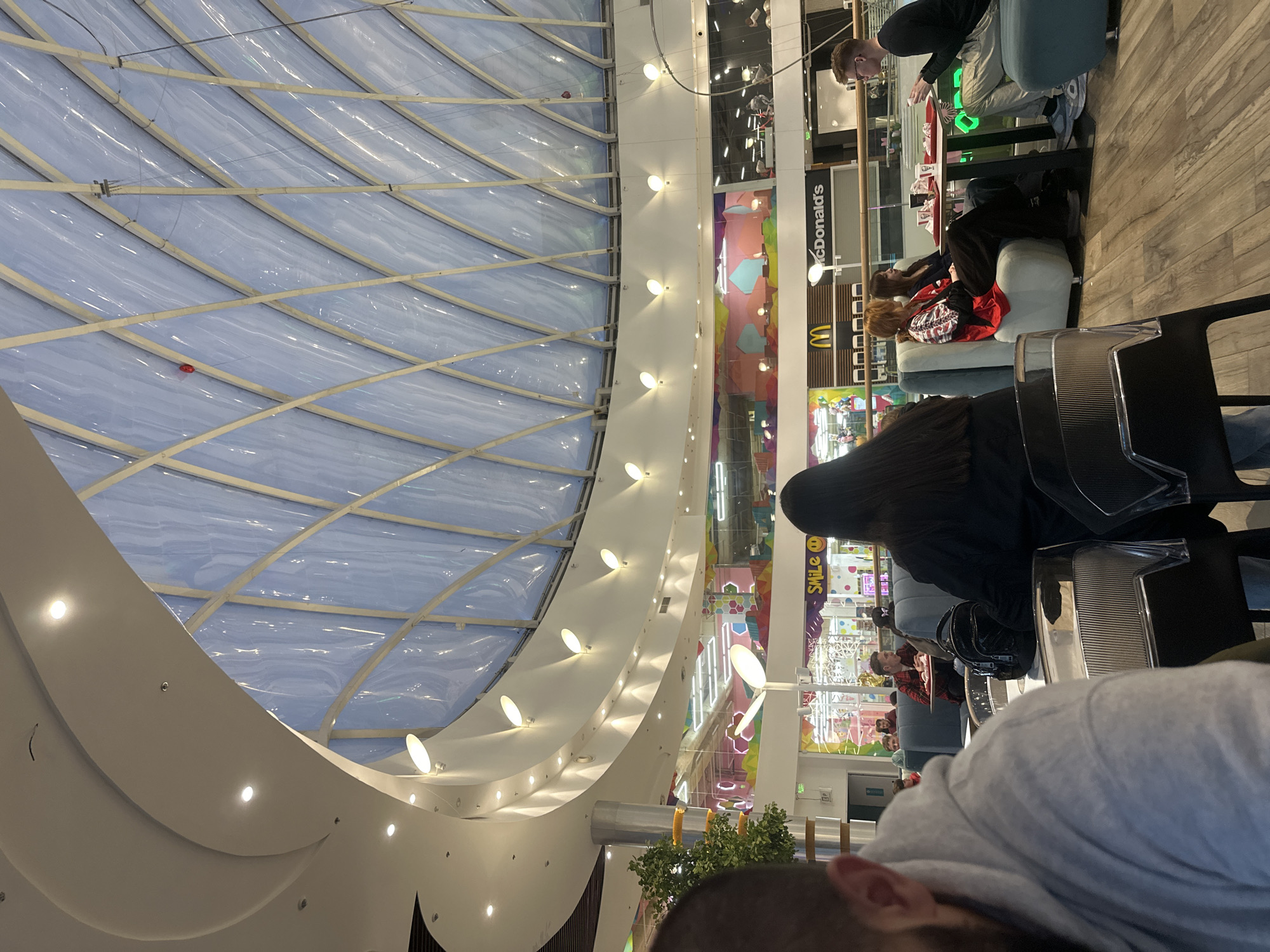 The image shows an interior view of a shopping mall in Kharkiv, Ukraine. The architectural design features a large, arched ceiling with a visible frame and translucent panels. Recessed lighting is spaced along the curved structure. Multiple shops and eateries line the mall's walkways, including a McDonald's restaurant. Several people are seated at tables and on lounge furniture, some appearing to be eating or conversing. The photograph is taken from the perspective of someone seated at a table, with the backs of two individuals visible in the foreground.