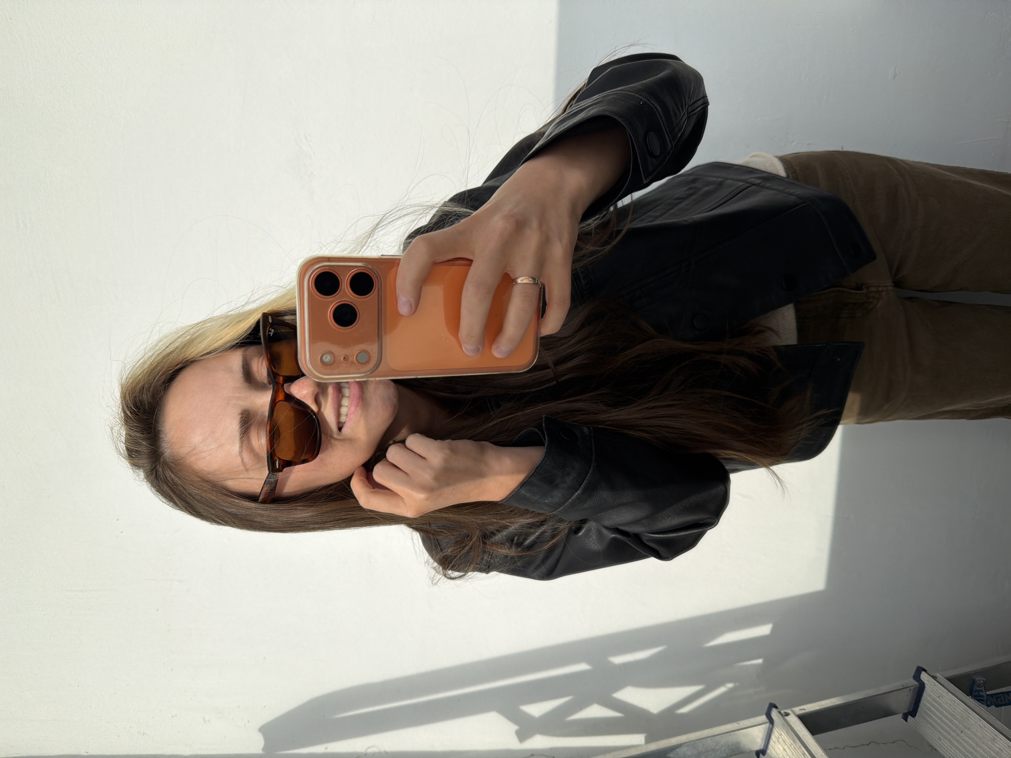 The image shows a young woman taking a selfie in a brightly lit indoor space with white walls and a floor that appears to be a light-colored material, possibly concrete. The woman has long, light brown hair and is wearing dark sunglasses and a black jacket over a light-colored top. She is also wearing brown pants. Her right hand is holding an orange-cased smartphone, angled to capture her reflection. She is smiling, with her teeth visible. A thin ring is visible on her left ring finger. Shadows are cast on the wall behind her, suggesting a light source, possibly a window, to her left. A ladder is visible in the lower right corner of the image, with some text on it that is partially obscured. The overall scene is casual and informal, likely a personal moment captured by the woman herself. The lighting suggests it is daytime. There are no explicit clues indicating the location is Khimki, Russia, beyond the prompt.