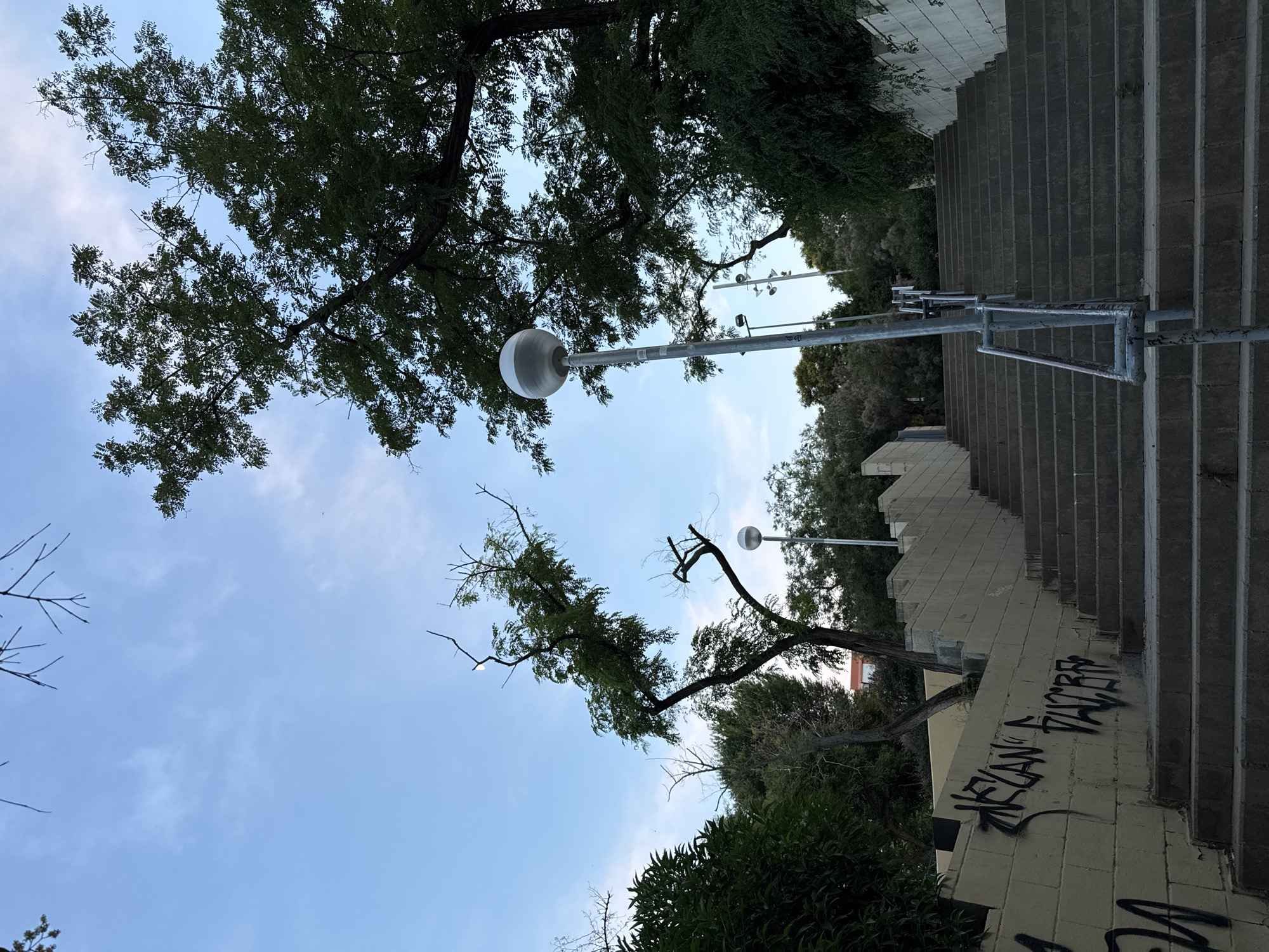 The image depicts an outdoor public stairway with a concrete surface, flanked by a yellow, graffiti-covered wall on the left and a white brick wall on the right. A metal handrail runs along the right side of the stairs. A modern lamppost with a spherical light fixture stands to the right of the stairway. Lush green trees and foliage surround the area, with a clear blue sky with sparse clouds overhead. The overall scene suggests a park or a well-trafficked pedestrian area in a Mediterranean climate. The presence of graffiti on the wall indicates an urban setting. The architecture and vegetation are consistent with locations in Barcelona, Spain.