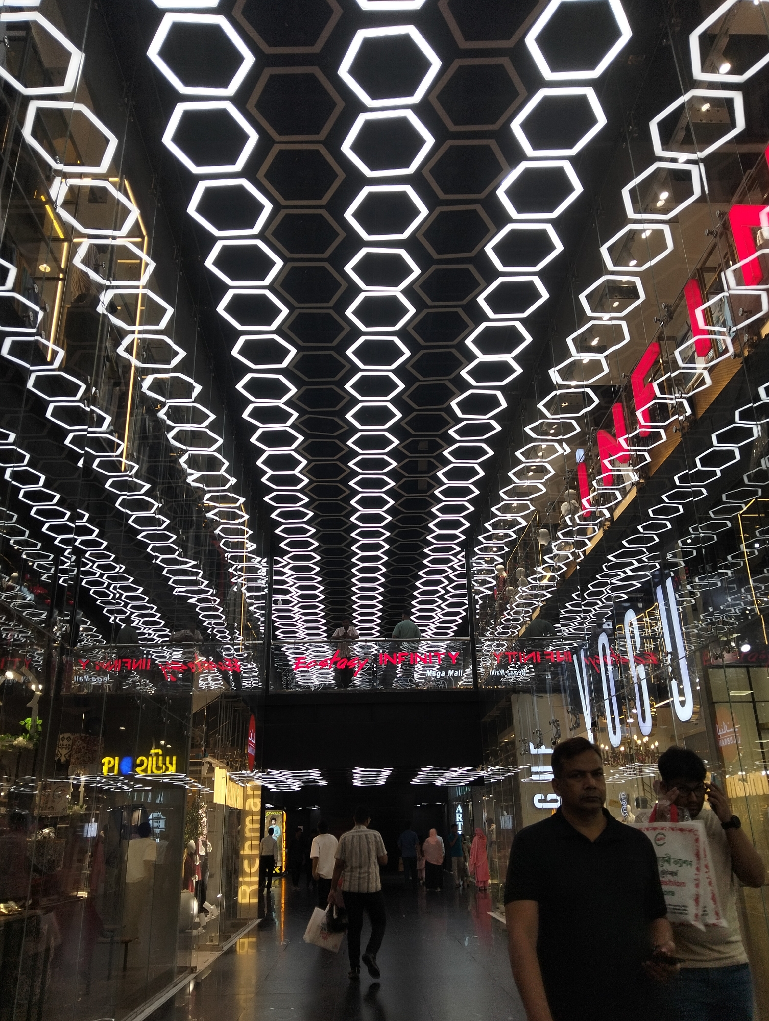 The image depicts an interior view of a shopping mall in Dhaka, Bangladesh. The ceiling is adorned with a pattern of hexagonal lights, creating a geometric effect. The walls are lined with storefronts, visible through glass. Several people are walking through the mall. Two men are in the foreground. One is wearing a black t-shirt, and the other is carrying a shopping bag and holding a phone to his ear. The mall appears to be well-lit and modern. Some storefronts display signs with writing in English and another language.