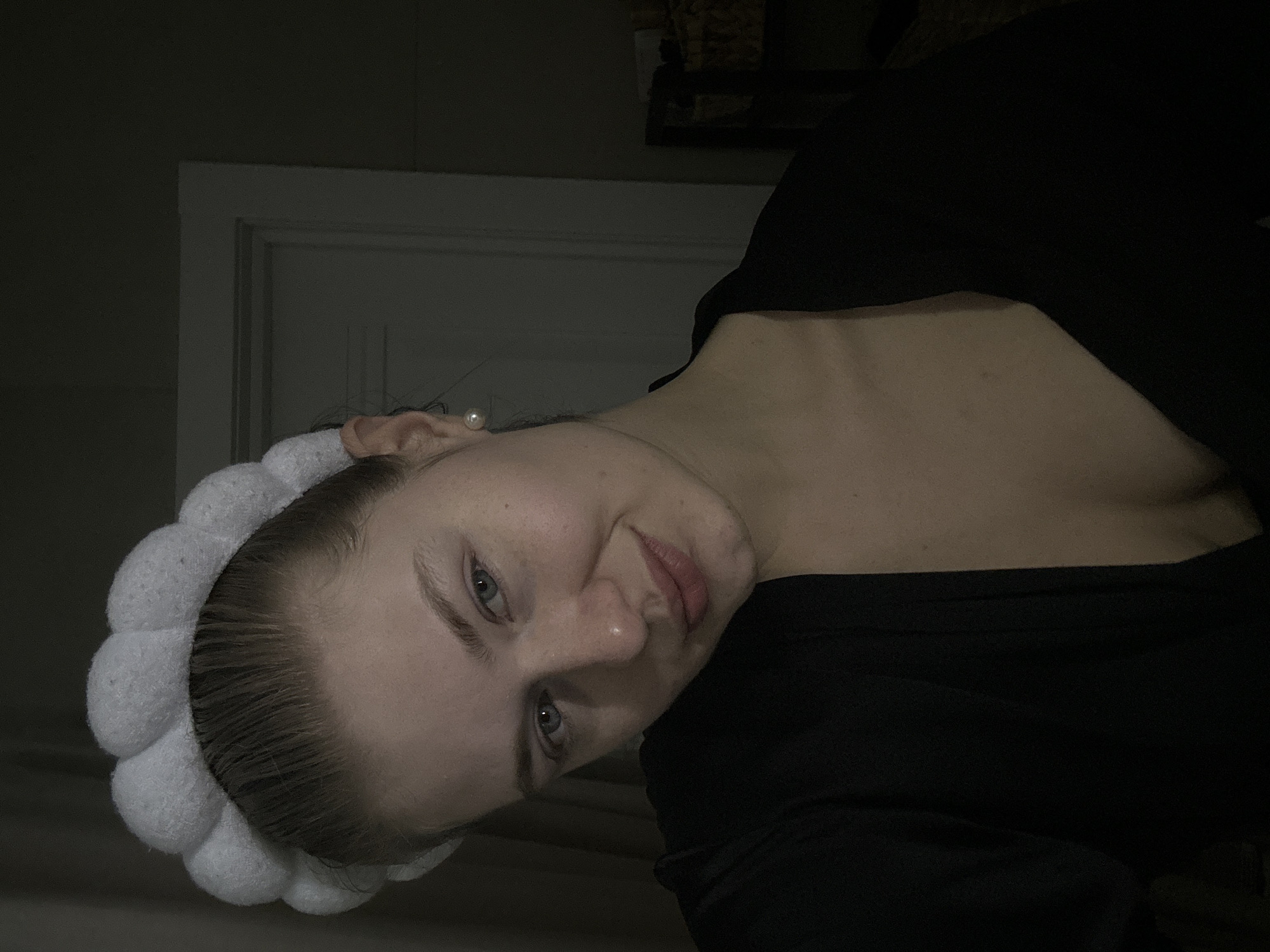 A single female subject is depicted in a dimly lit indoor environment, stated to be in Kazan, Russia. The individual has fair skin, blue eyes, and light hair pulled back, secured by a white, segmented, puffy headband. A small, round, white pearl stud earring is visible in her right ear. She wears a dark-colored, V-neck garment, revealing a portion of her upper chest. The subject is looking directly at the camera with a slight smile. The background on the right includes a partial view of a white doorframe and a segment of a wicker basket. The left side of the background is indistinct and dark, possibly a curtain or wall.
