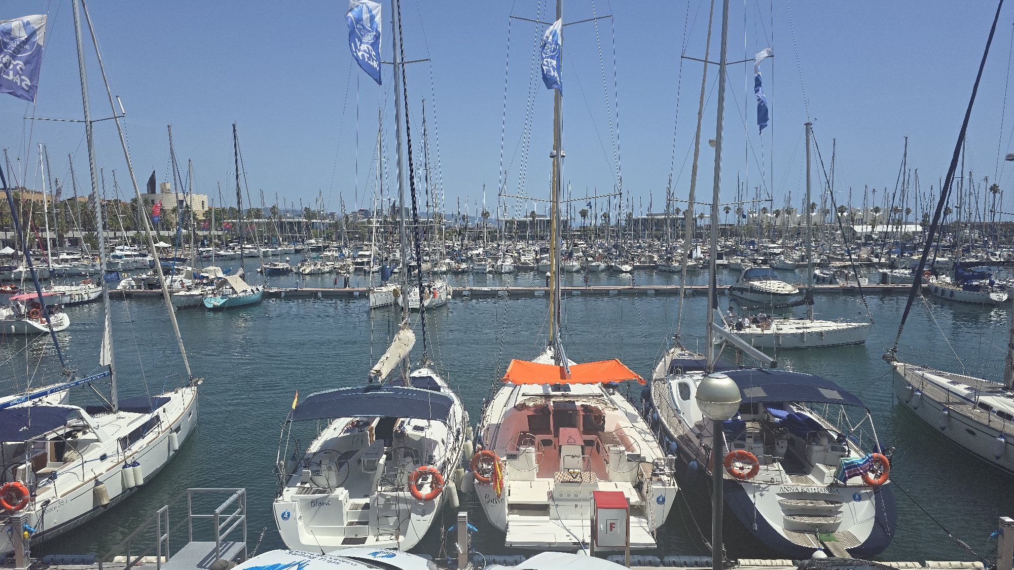 The image depicts a large marina filled with numerous sailboats moored in the Port Olímpic of Barcelona, Spain. The scene is bathed in bright sunlight under a clear blue sky. In the foreground, three white sailboats are prominently featured, side-by-side, with their sterns facing the viewer. The leftmost boat has a blue canopy and a life preserver attached to its railing. The middle boat is adorned with an orange tarp stretched over its cockpit and has a Spanish flag flying from its stern. The rightmost boat also has a blue canopy and the name "Blues" is visible on its stern. Behind these boats, a vast expanse of similar vessels fills the harbor, with masts creating a dense forest-like appearance. In the background, the city skyline of Barcelona rises, with buildings and palm trees visible, indicating an urban coastal environment. The water is calm and reflects the sky and the boats. Two blue flags with white lettering are visible on tall poles, fluttering in the breeze, one on the left and another towards the center of the image. The overall impression is one of a busy, well-maintained nautical hub.