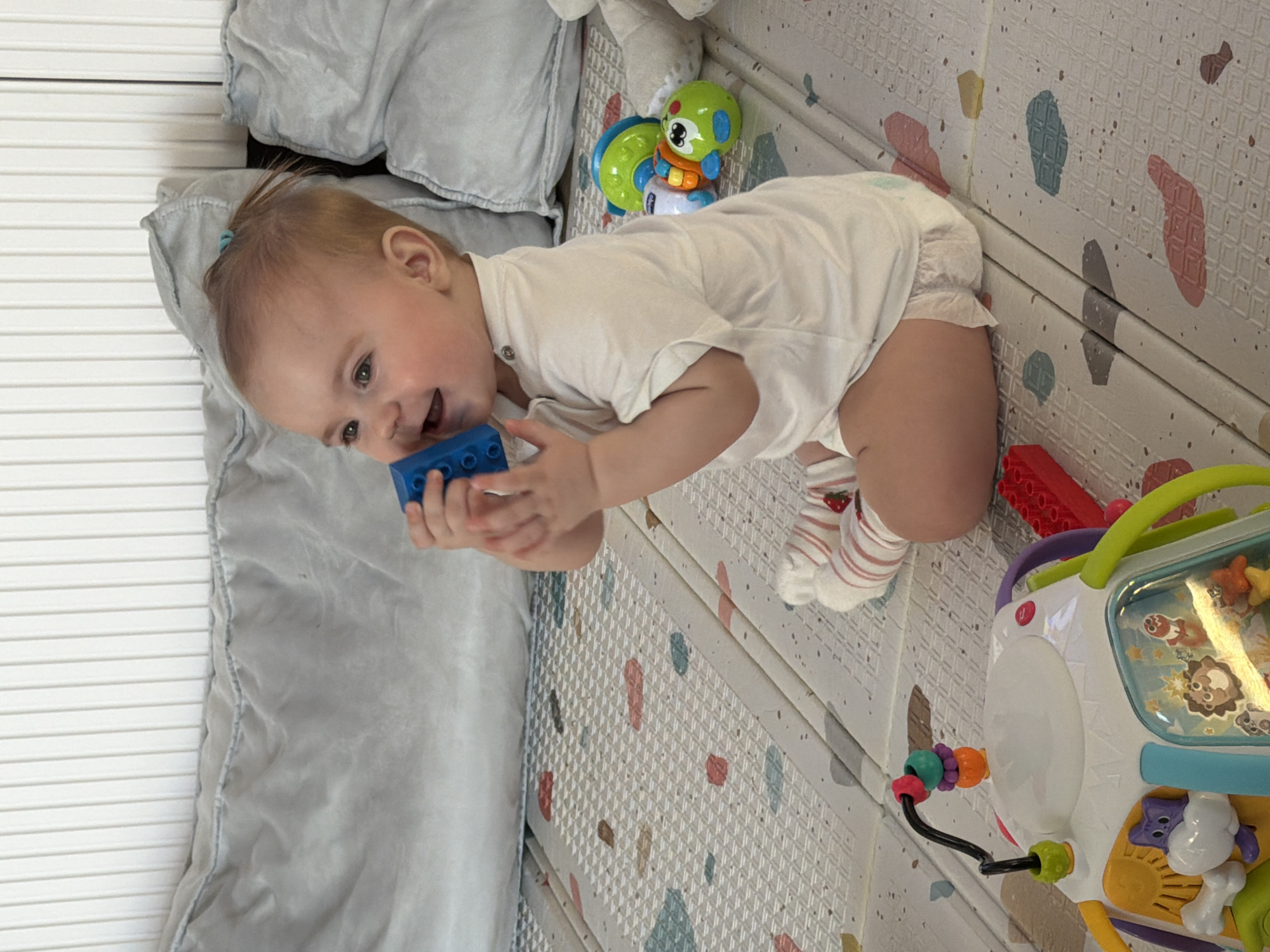 A baby is kneeling on a light-colored playmat with colorful shapes, holding a blue block. The baby is wearing a white shirt, a diaper, and striped socks. To the left of the baby is a gray quilted crib bumper next to white slatted crib sides. Behind the baby are additional toys, including a green and blue toy, a red toy, and a white activity center. The location is a home setting, and there is no specific evidence in the image to indicate it is in Krasnoyarsk, Russia.