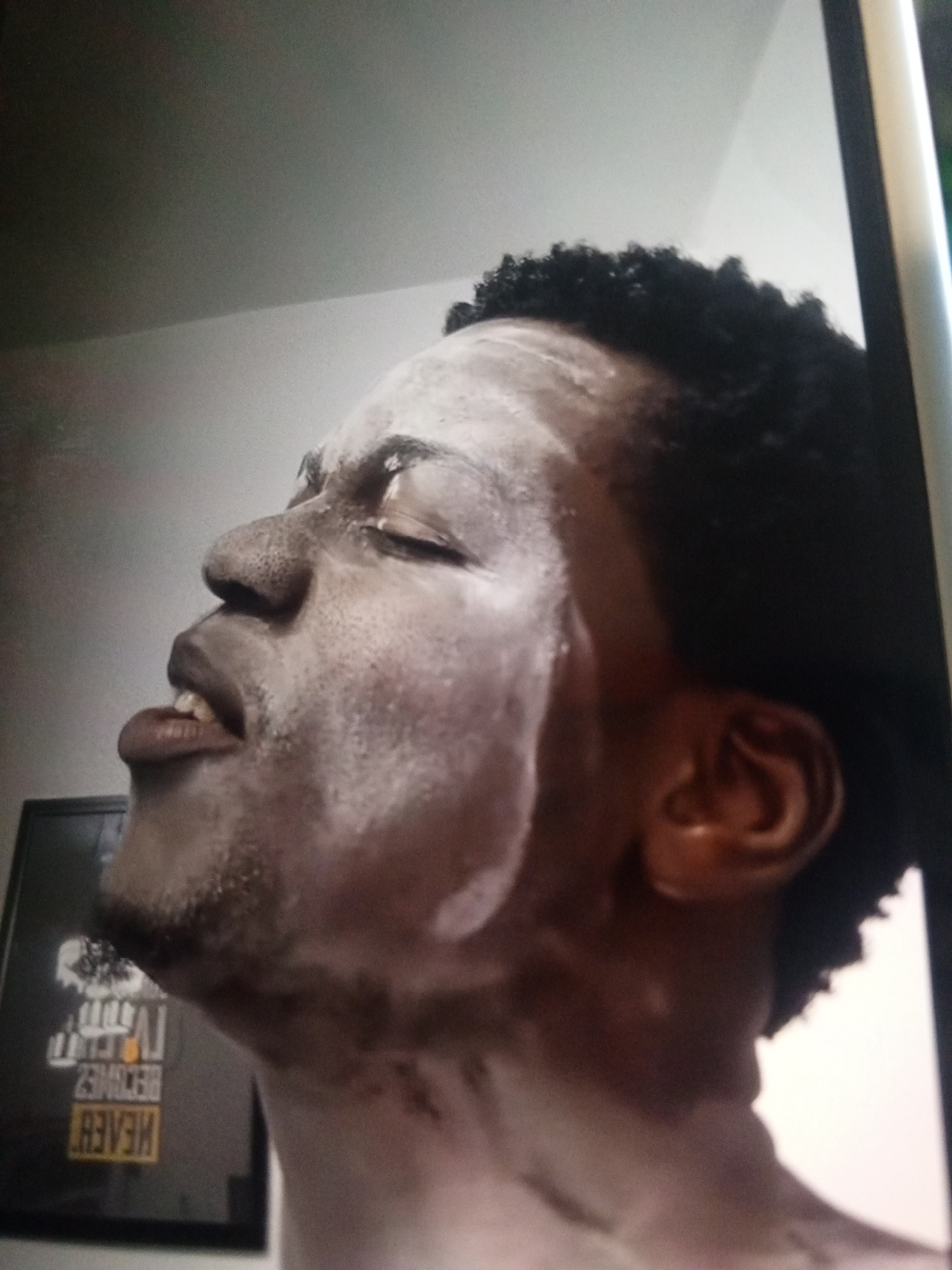 A close-up, upward angle shot of a man's face covered in a white substance, possibly a facial mask. He has dark, curly hair, closed eyes, and his mouth is slightly open. His head is tilted back. In the background, there is a wall and a framed picture with yellow writing. The specific location within Unknown, Unknown is indiscernible from the image.