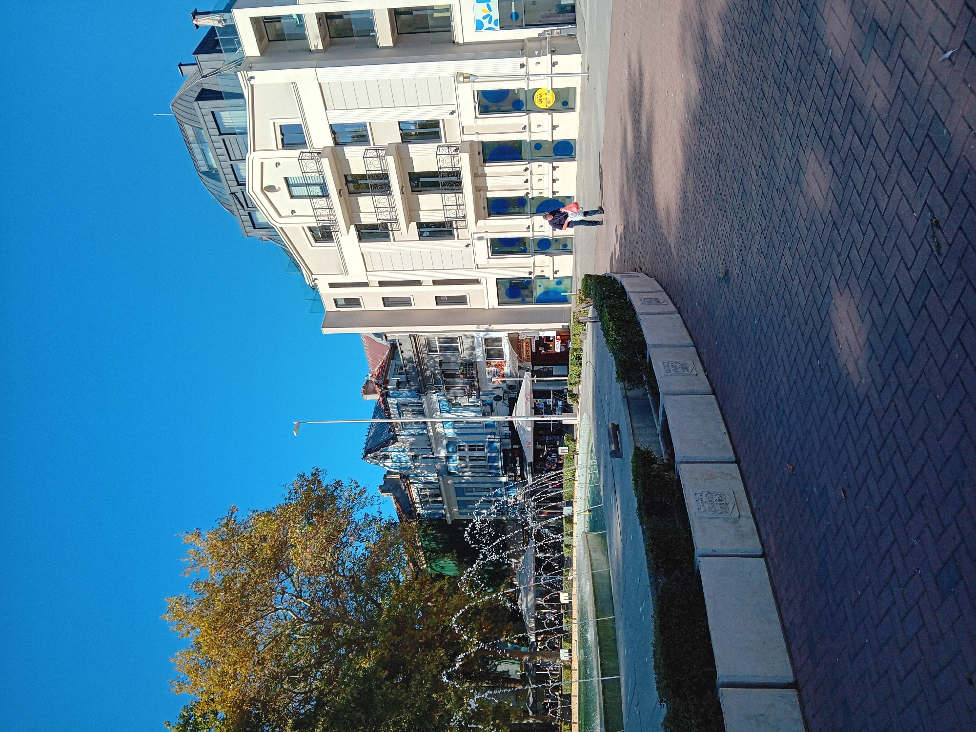 The image captures a view of the pedestrian area in front of the Municipality of Varna, Bulgaria, with a focus on a public fountain in the foreground. The fountain's water jets are actively spraying, creating a dynamic element in the scene. Beyond the fountain, a paved walkway leads towards a row of buildings.
To the left, a large tree with autumn foliage provides shade over outdoor seating areas, likely belonging to cafes or restaurants. The buildings in the background exhibit a mix of architectural styles. One prominent structure features a modern facade with balconies and a curved, metallic roof. Adjacent to it, older buildings display traditional European architecture with blue and white detailing.
A single individual is visible walking on the pavement, carrying a red bag. The ground is paved with dark red interlocking bricks laid in a herringbone pattern. The sky is a clear, bright blue, suggesting fair weather. The overall scene depicts a well-maintained urban public space, likely a popular gathering spot in the city.