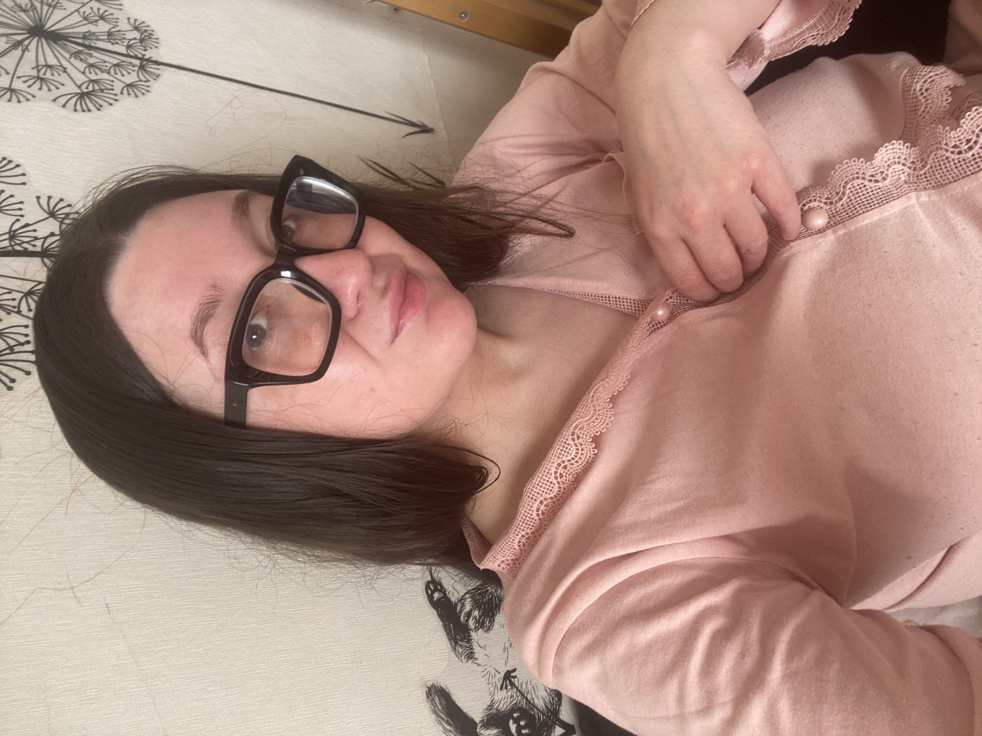 The image displays an individual, identified as female, positioned indoors within the city of Chaikovsky, Russia. She is presented in a chest-up portrait, looking directly forward with a subtle expression. The subject has long, dark hair and wears thick-framed black eyeglasses. She is dressed in a light pink, long-sleeved top that features lace trim along the V-neckline and cuffs. Pearlescent pink buttons are visible down the front of the garment, with her left hand resting on the fabric near these buttons. The background consists of a light-toned wallpaper adorned with dark patterns, specifically stylized dandelion seed heads visible on the upper right, and a partial illustration of a stylized rabbit figure on the left. Faint red and blue marks, along with a black arrow-like drawing, are present on the wallpaper. A segment of a light-colored wooden structure is visible along the far right edge of the frame.