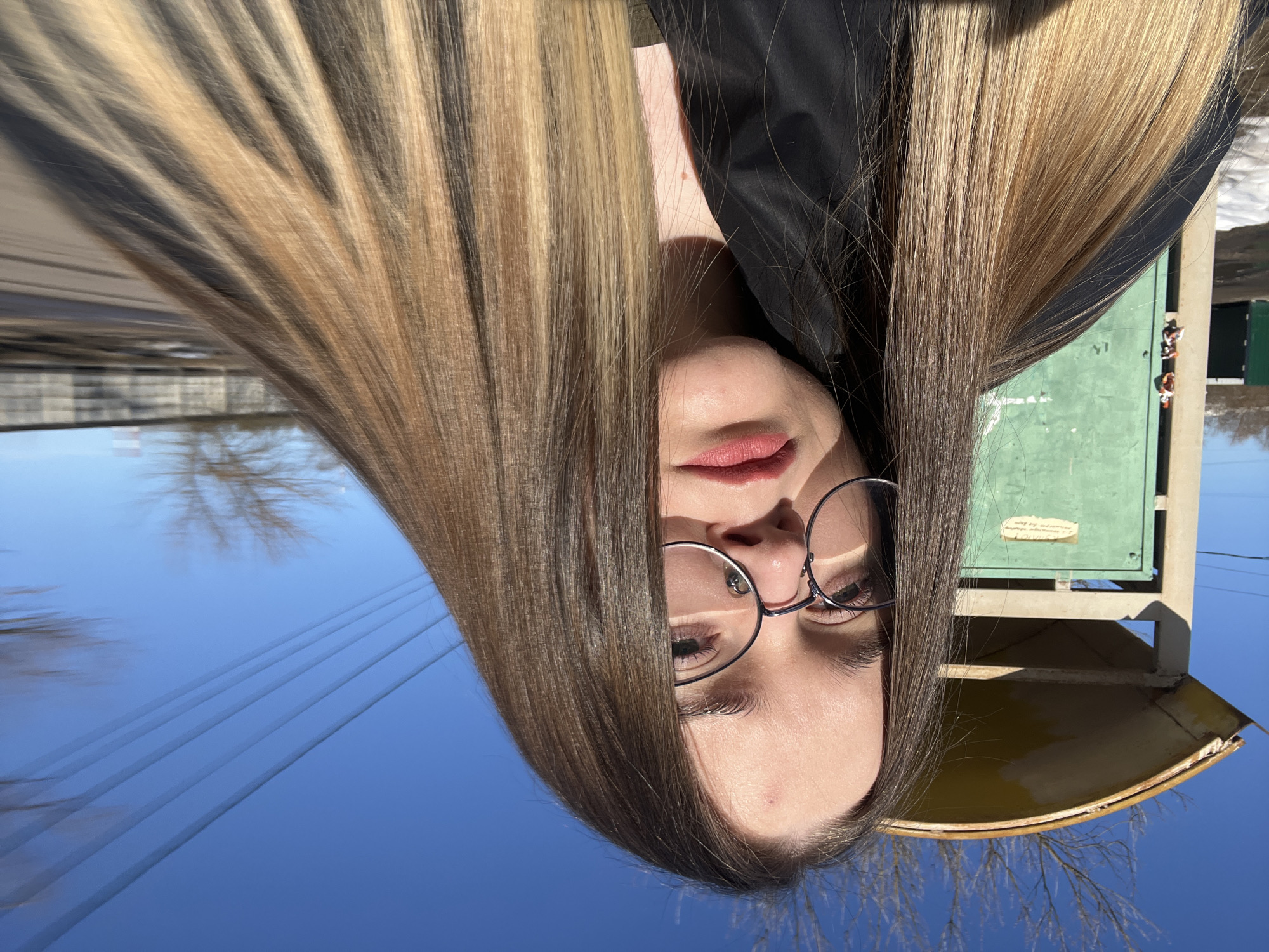 A young woman with long, straight, dark brown to blonde ombre hair, wearing round metal-frame glasses and dark clothing, is centered in a selfie, looking directly at the camera. Her lips are adorned with red lipstick, and a small blemish is visible on her forehead. To her left, partially obscured, is a weathered structure featuring a yellow, curved corrugated metal roof atop a rectangular, green-painted panel, resembling a bus stop or small shelter. A white paper is affixed to the green panel. Bare tree branches are visible above this structure against a clear blue sky. Multiple utility lines are discernible in the upper right quadrant. Patches of snow lie on the ground in the lower left, indicating cold conditions. In the blurred background to the right, additional bare trees, a grey structure consistent with a fence or building, and a road or dirt path are present. This outdoor scene is located in the city of Большевик, Russia.