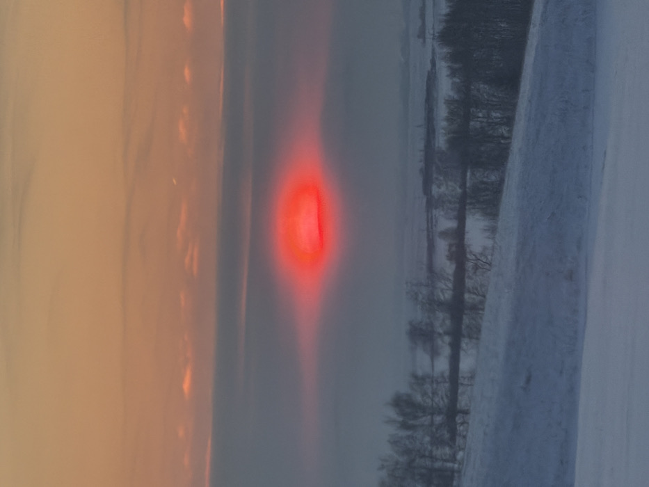 A landscape portrait shows a vibrant red glow piercing a foggy, possibly winter, scene. To the right, snow-covered ground slopes upward with bare trees standing against the soft, muted backdrop. Left of the red phenomenon is an orange-tinted sky transitioning into the hazy middle ground where the red light dominates the scene. The orientation is horizontal, however, the landscape has been rotated clockwise.