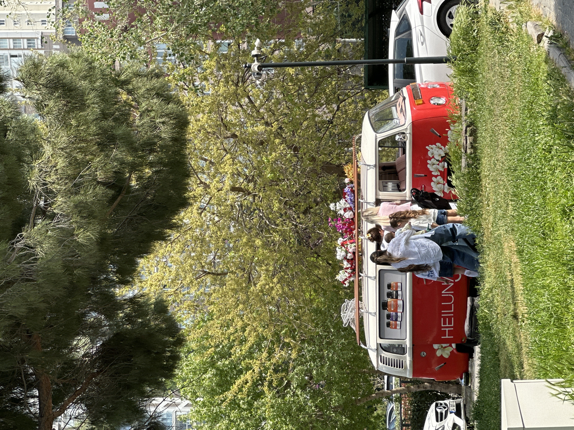 The image shows a red and white Volkswagen bus with floral decorations parked on a grassy area. The word "HEILUNA" is visible on the side of the bus. Several people are interacting around the bus, with some possibly looking at or purchasing items from it. The environment is green with trees in the background and buildings in the distance. The scene appears to be in an urban setting, possibly a park or open area within the city of Başakşehir, Turkey.
