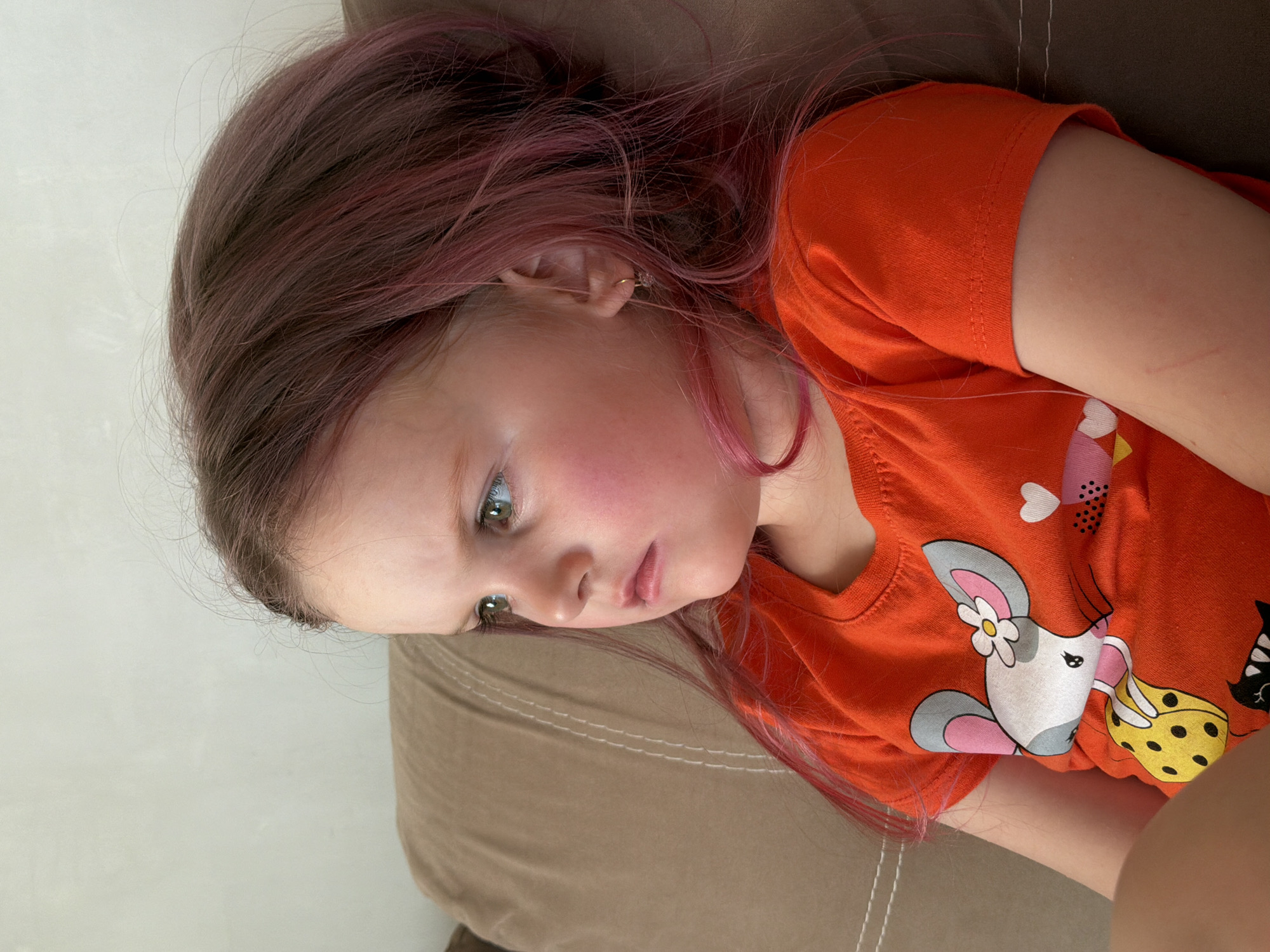 A young child, likely a girl, is depicted in a high-angle, close-up shot, appearing to lie down or recline indoors. She has long, straight hair that is primarily brown with prominent pink streaks or highlights, especially visible towards the ends and around her face. Her light-colored eyes, appearing green or hazel, are cast downwards and slightly to the left, giving her a pensive or contemplative expression. Her cheeks are noticeably flushed or rosy.
She is wearing a vibrant orange short-sleeved t-shirt adorned with a colorful cartoon graphic. The most prominent part of the graphic features a stylized grey and pink mouse-like creature with large ears and a white flower tucked behind one ear. This character is shown holding a yellow, black-polka-dotted object, possibly cheese. Other small, illustrative elements like hearts and a dark, possibly cat-like figure, are also visible on the shirt. A small gold hoop earring with a delicate, light-colored bead or gemstone is visible in her left ear.
The child rests on a brown upholstered surface, which could be a sofa or a large pillow, with a plain, light off-white background behind her. Several faint red linear scratches are visible on her left forearm, just above the cuff of her t-shirt sleeve. The scene suggests a quiet, candid moment of rest or observation in an indoor home setting. No text is visible in the image, and there are no direct visual cues for the time of day.
