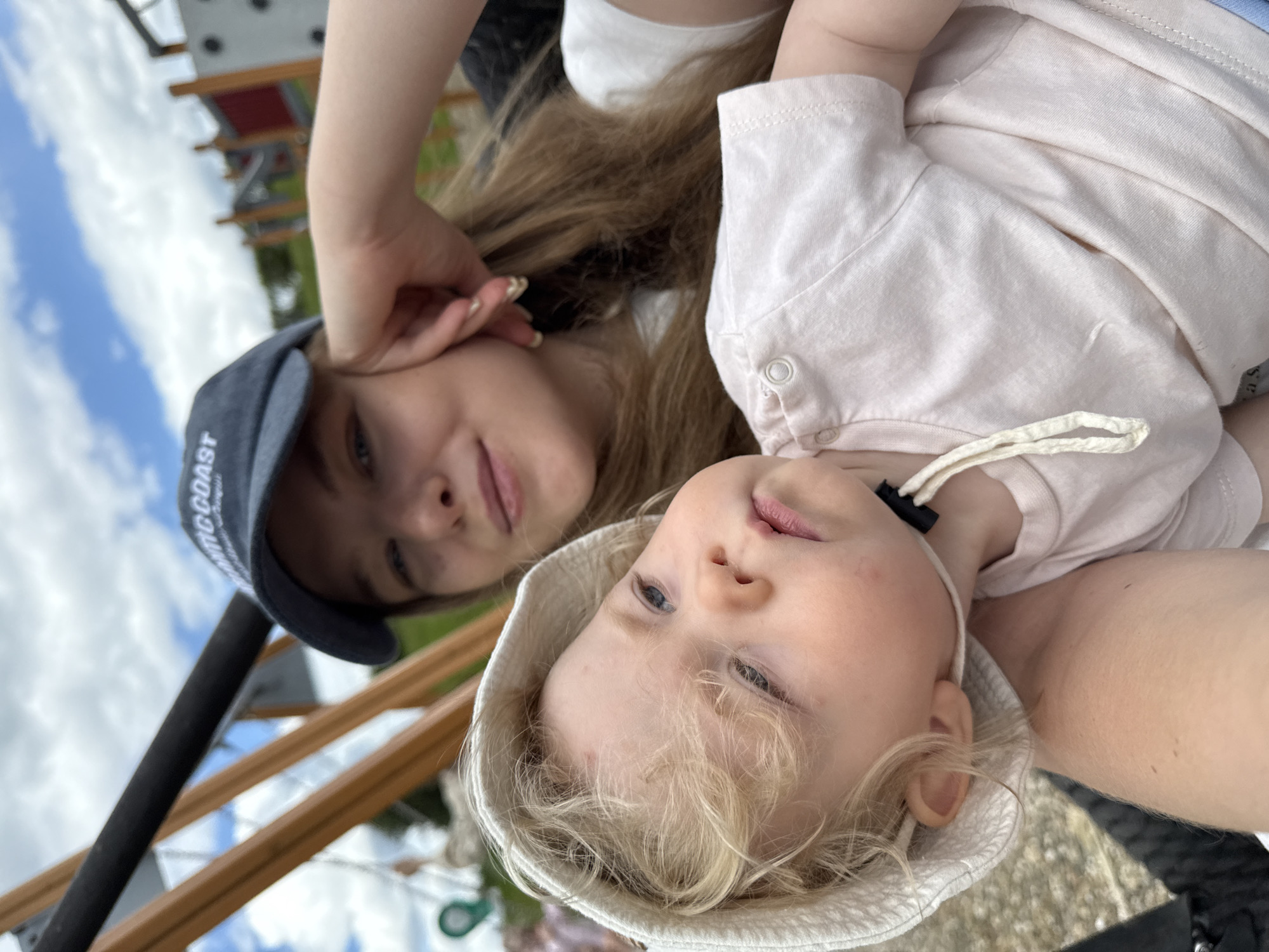 This outdoor selfie-style image, rotated counter-clockwise, features an adult female and a young child. The sky, partially cloudy with blue patches, appears on the left side of the frame, indicating a bright daytime setting.
The adult, positioned slightly behind and above the child, has long, light brown hair and fair skin. She wears a dark blue baseball cap, on which the text "MINGCOAST" is visible in white letters. Her face, from the nose up, is visible, showing a calm, relaxed expression with a hint of a smile, and one hand is raised towards her ear.
In the foreground, a young child, likely a toddler, is visible with light blonde, curly hair and fair skin. The child wears a light-colored, possibly white or very light beige, bucket hat and a light pink or beige long-sleeved top. The child's face shows a calm, curious expression, looking slightly towards the camera.
The background reveals wooden structures, which appear to be railings or support beams, possibly part of a playground, an observation deck, or a similar outdoor recreational facility. A small section of a distant structure with a red roof element can also be seen. The scene depicts a casual, pleasant interaction between the two individuals in an outdoor setting in Dobrograd, Russia.