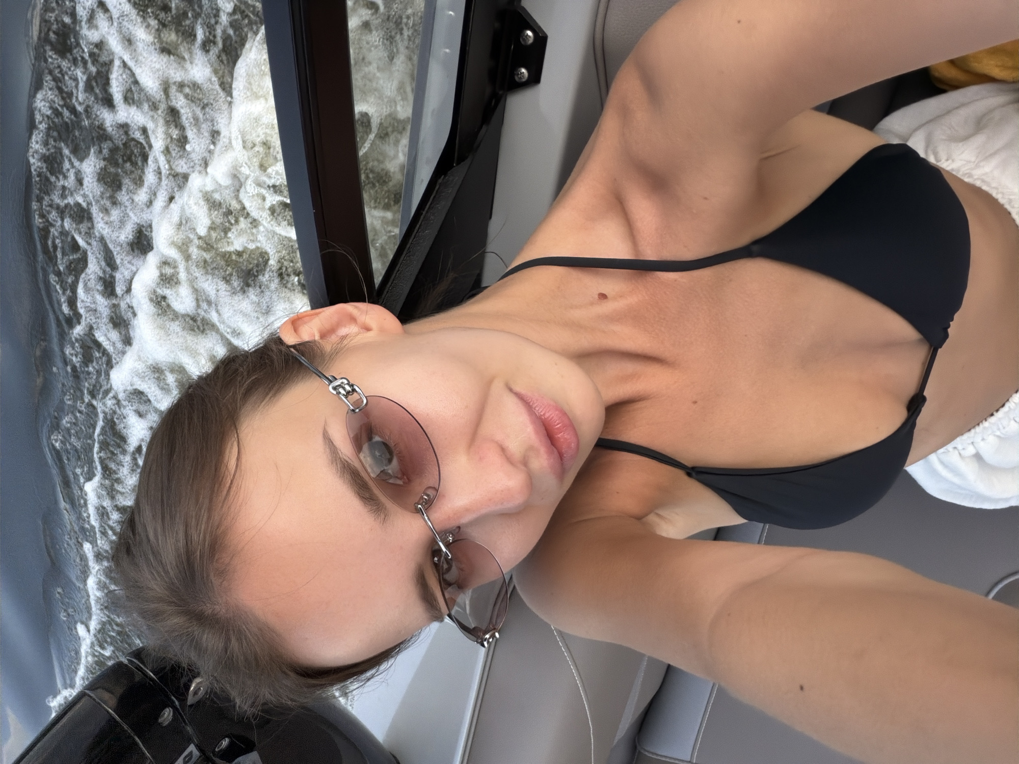 A young woman with brown hair pulled back, wearing sunglasses and a black bikini top, is taking a selfie on a boat. The water behind her is choppy with white foam, suggesting the boat is moving. The background shows the dark railing of the boat and some of the seating. The image appears to be taken during the daytime. The location is identified as Terekhovo, Russia, which is a village located on the outskirts of Moscow, known for its recreational areas and proximity to the Moskva River.