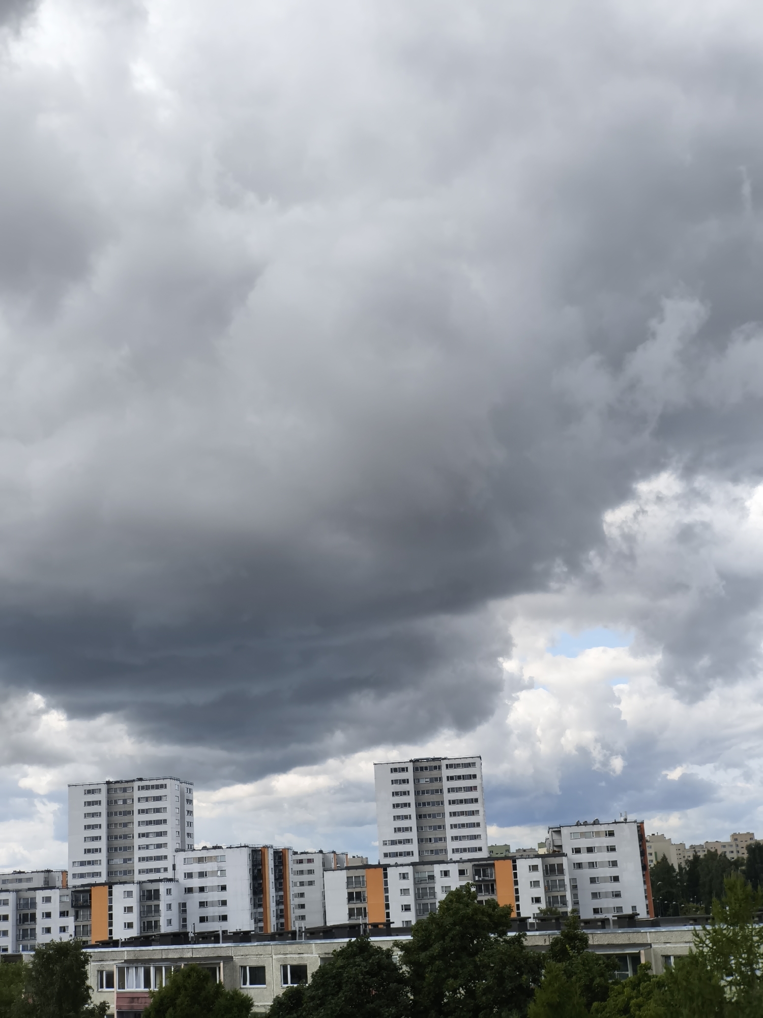 The image depicts a cloudy sky over an urban landscape in Tallinn, Estonia. In the foreground, several multi-story residential buildings are visible. These buildings are predominantly white with some sections featuring orange accents and dark gray trim. The architecture suggests a post-Soviet era design, common in many parts of Tallinn. Lush green trees and foliage partially obscure the lower portions of some buildings. The sky is filled with heavy, dark gray storm clouds, with only small patches of lighter clouds and a hint of blue sky visible towards the right side of the frame. The lighting is diffused, typical of overcast conditions. No people are discernible in the image. The scene conveys a sense of impending weather change.