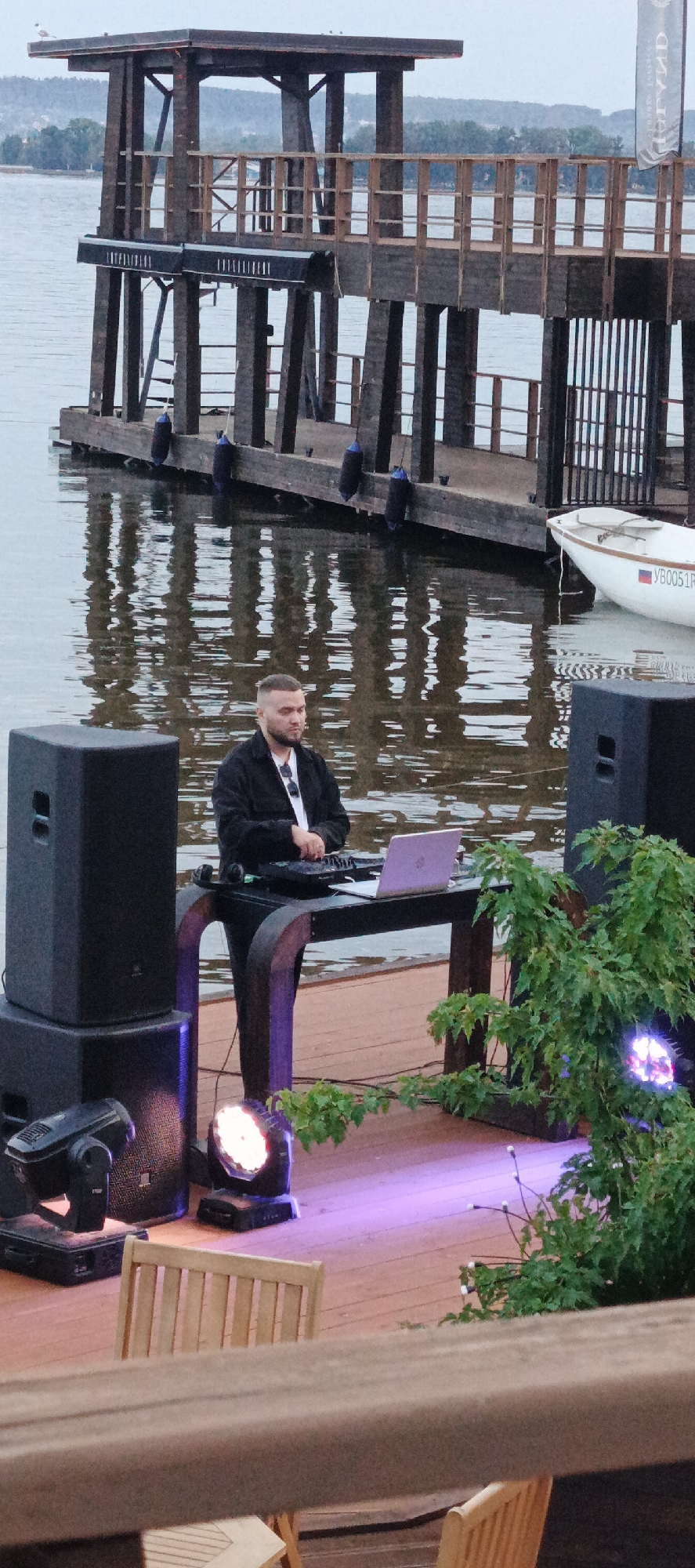 The image depicts an outdoor scene near a body of water, likely a lake or river. In the foreground, a DJ, a male with short dark hair, is positioned behind a table with a laptop and DJ equipment. He is wearing a black jacket over a white shirt and sunglasses are tucked into his jacket. Two large black speakers are placed to his left, and a stage light is visible on the wooden platform in front of him, casting a purple glow. To the right of the DJ, lush green foliage and another stage light are present. In the background, a wooden structure resembling a pier or boardwalk with railings extends over the water. A small white boat with registration numbers "YB0051R" is docked to the right of the wooden structure. The water reflects the wooden structure and the sky. The overall setting suggests an event taking place on a waterfront. The provided context mentions Votkinsk, Russia, which is known for its picturesque location on the Votkinsk Reservoir, part of the Kama River system. This location is consistent with the visual elements in the image, particularly the presence of a large body of water and a wooden pier or boardwalk.