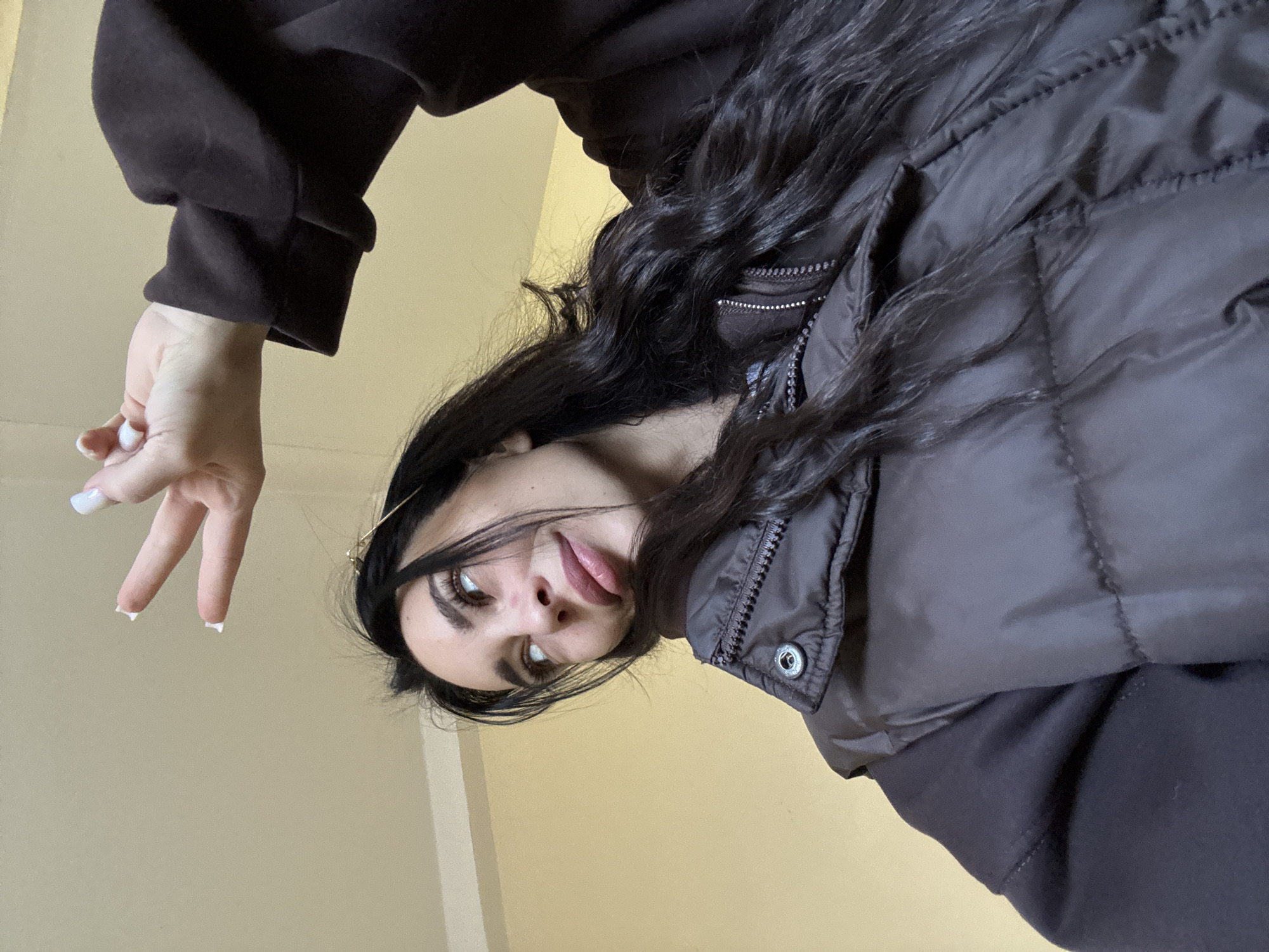 A woman with dark hair is looking down at the camera with a slight smile. She is wearing a dark brown vest over a brown hoodie. Her left hand is raised in a peace sign. The background appears to be a plain wall. There is no information in the image to indicate it was taken in Chaikovsky, Russia.