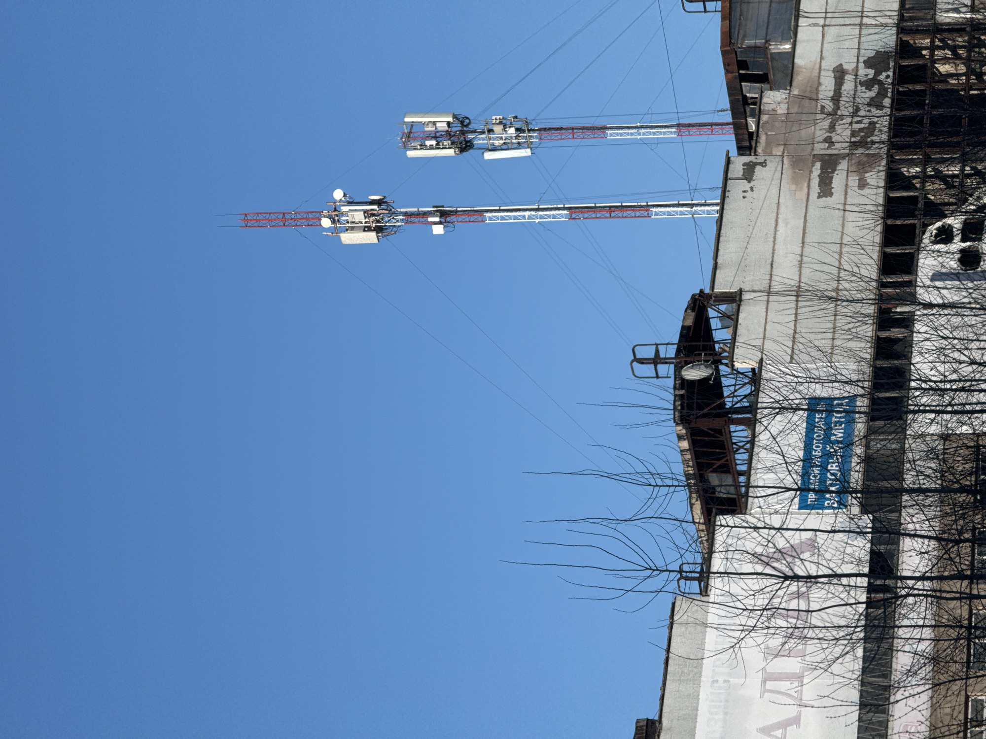 The image shows a view of a building in Chaikovsky, Russia. The building appears to be multi-storied and has a large faded advertisement on its side. The building has metal scaffolding on the side. Two cell phone towers are attached to the building's roof. The sky is clear and blue. The trees in the foreground are bare of leaves. The location is in Chaikovsky, Russia, as indicated by the Cyrillic text on the building.
