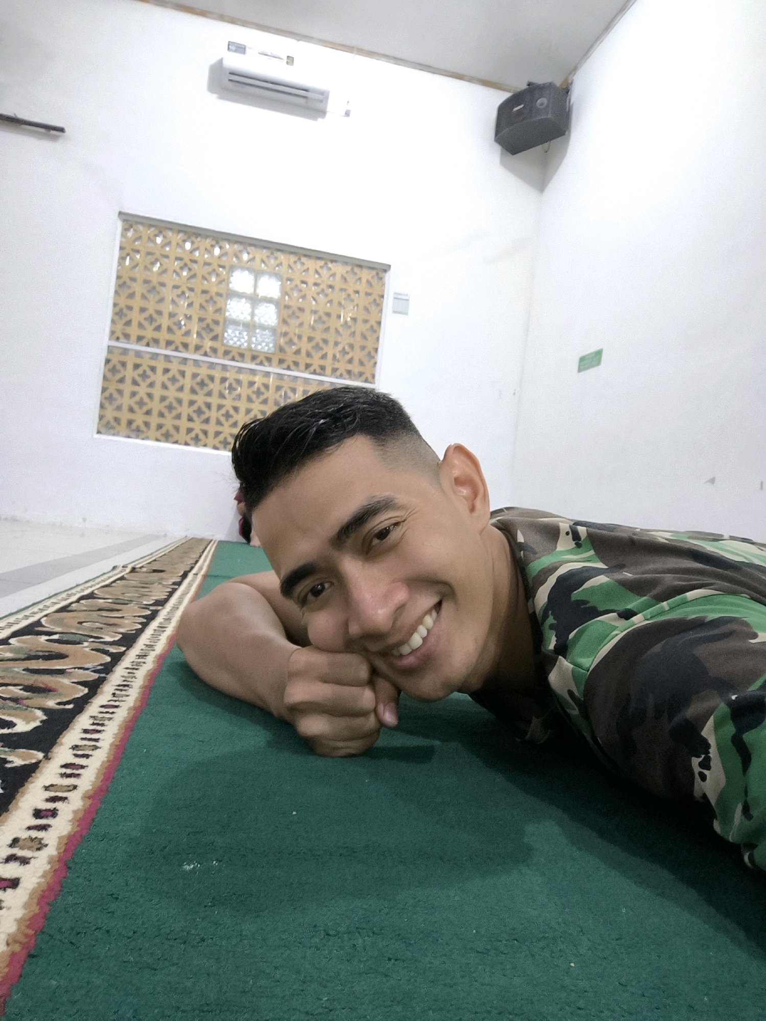 In an indoor setting, a male in camouflage clothing lies on a green carpet, resting his head on his hand. He smiles at the camera, with his body angled slightly away. A prayer rug with a decorative border runs along the left side of the frame, partially visible. The walls of the room are white, and a decorative window with a lattice pattern is visible in the background, along with an air conditioning unit mounted high on the wall. A black speaker is mounted in the corner of the ceiling. The overall environment suggests a room that could be a mosque or a common area within a facility. The lighting is bright and even, illuminating the scene clearly.