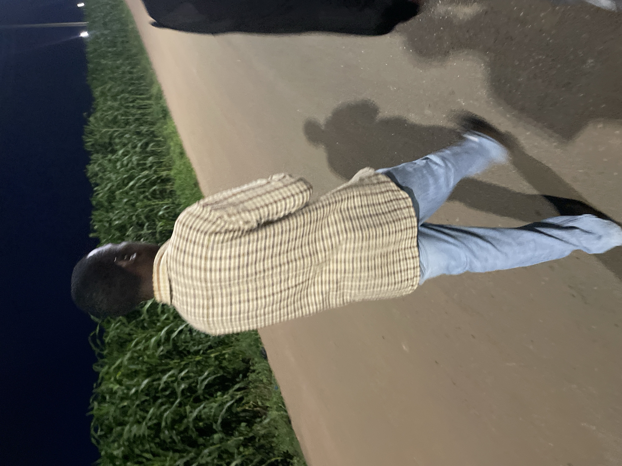 A person, viewed from behind, walks away from the camera on a dirt road at night in Barakin Lerawi, Nigeria. The individual wears a light-colored plaid shirt and blue jeans. To the left of the road is a dense field of green corn stalks. The sky above is dark and clear. The road is illuminated by artificial light, casting shadows on the ground. Another person, mostly out of frame, is visible to the right.