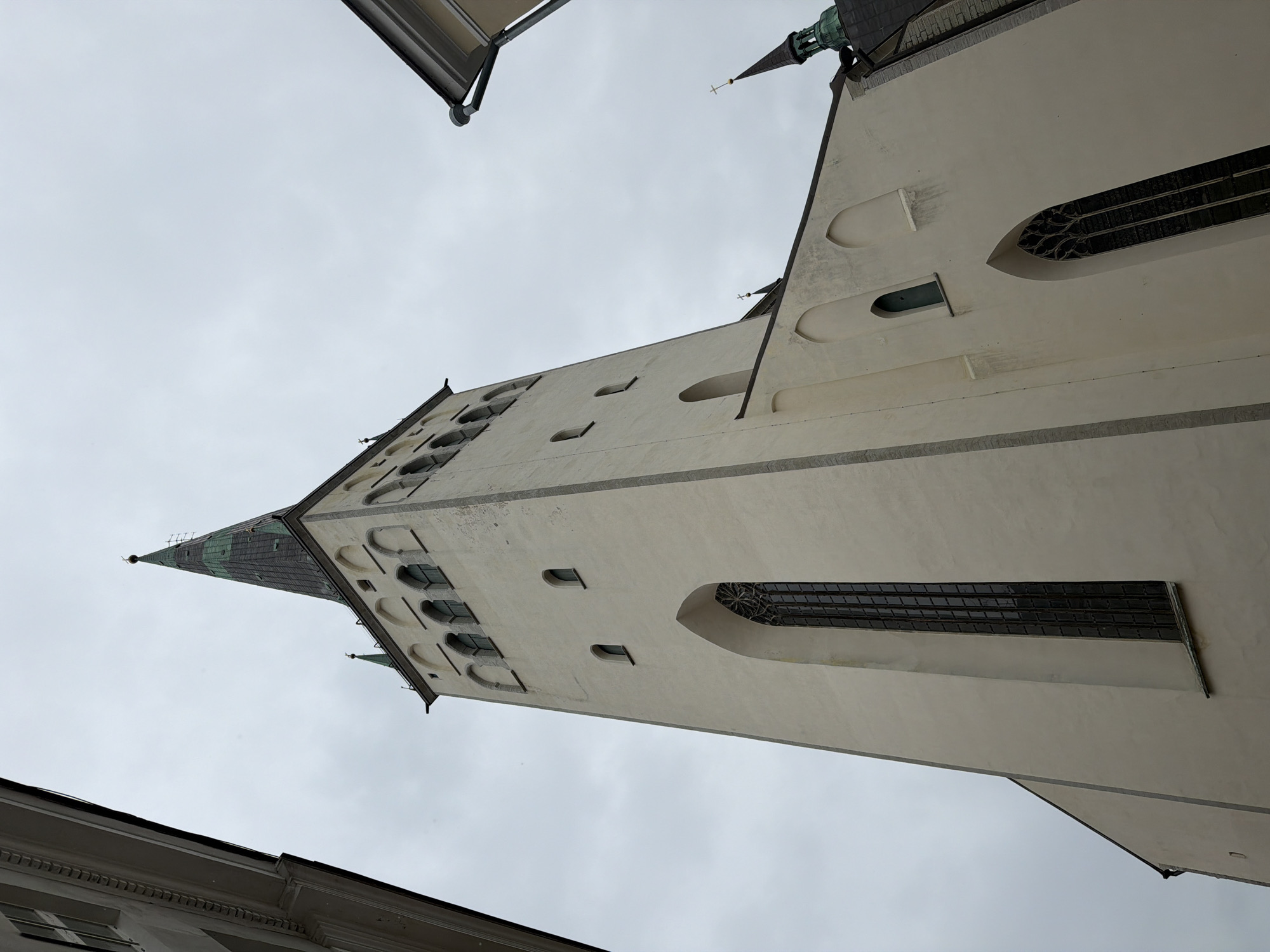 The image shows a low-angle view of the Oleviste Church (St. Olaf's Church) in Tallinn, Estonia, on an overcast day. The church is characterized by its tall, white facade, a prominent spire with a green roof, and numerous arched windows. Adjacent buildings partially frame the view of the church. The sky is a uniform gray, indicating cloudy weather. There are no people visible in the image. The architecture suggests a historical religious building with Gothic influences.