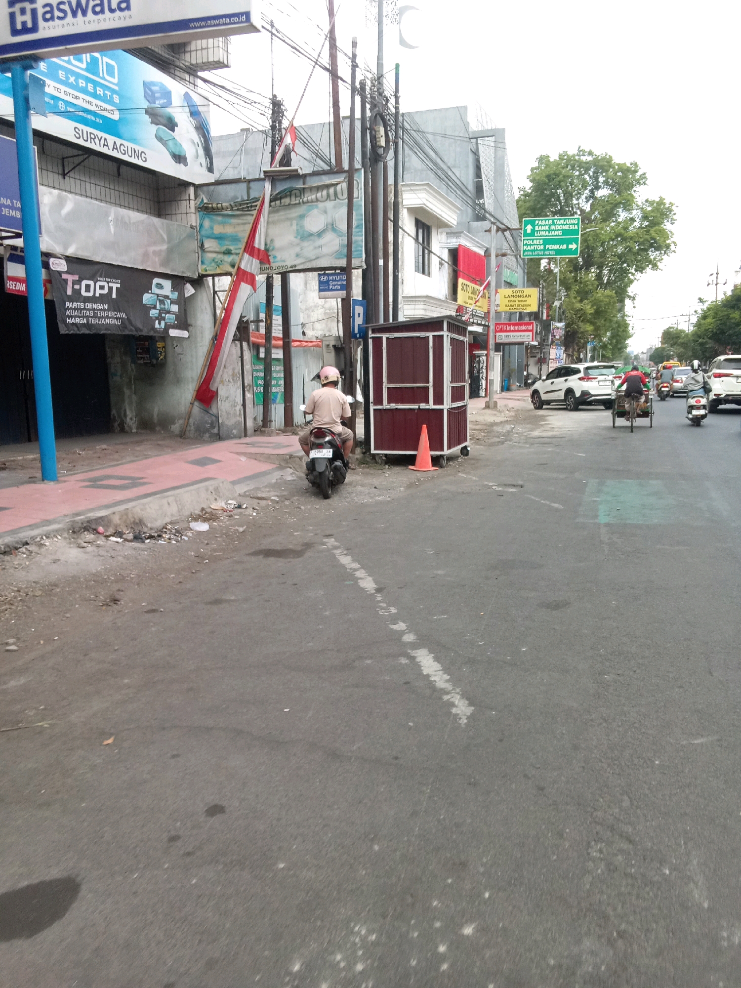 The image shows a street scene in Jember, Indonesia. On the left side of the road, there is a sidewalk with several advertisements. One large blue and white advertisement reads "EXPERT'S". Below this, a red and white Indonesian flag is displayed vertically, attached to a pole. Further down the sidewalk, there is a dark-colored kiosk with a red roof and a small orange traffic cone in front of it. A person wearing a light brown shirt and a pink helmet is riding a motorcycle away from the camera, moving along the right side of the street. The road is paved and appears to be a two-lane road with some traffic. In the background, there are buildings, trees, and more signs. One green directional sign indicates "PASAR TANJUNG BANK INDONESIA LUMAJANG". Another sign points to "POLRES KANTOR PEMKAB". Several cars and a bicycle rickshaw are visible on the road. The overall atmosphere is that of a busy street in an urban area.